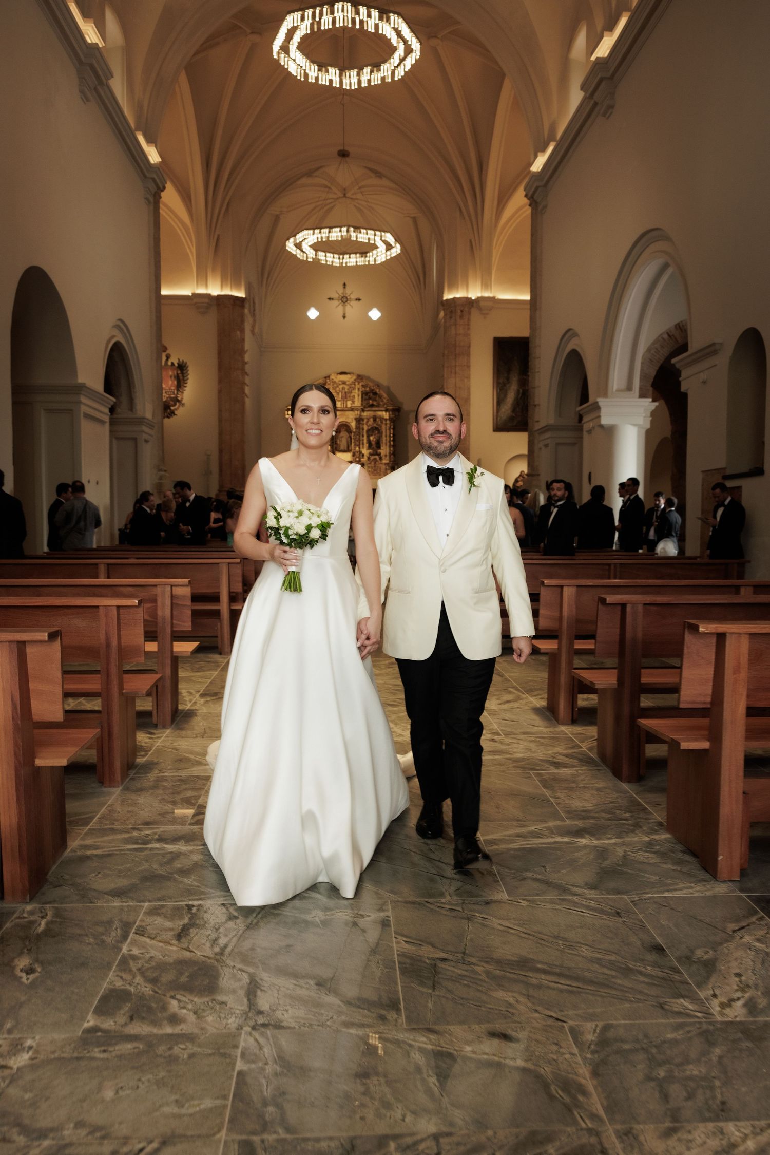 A wedding couple walks down the aisle of a church with vaulted ceilings and elegant chandeliers.