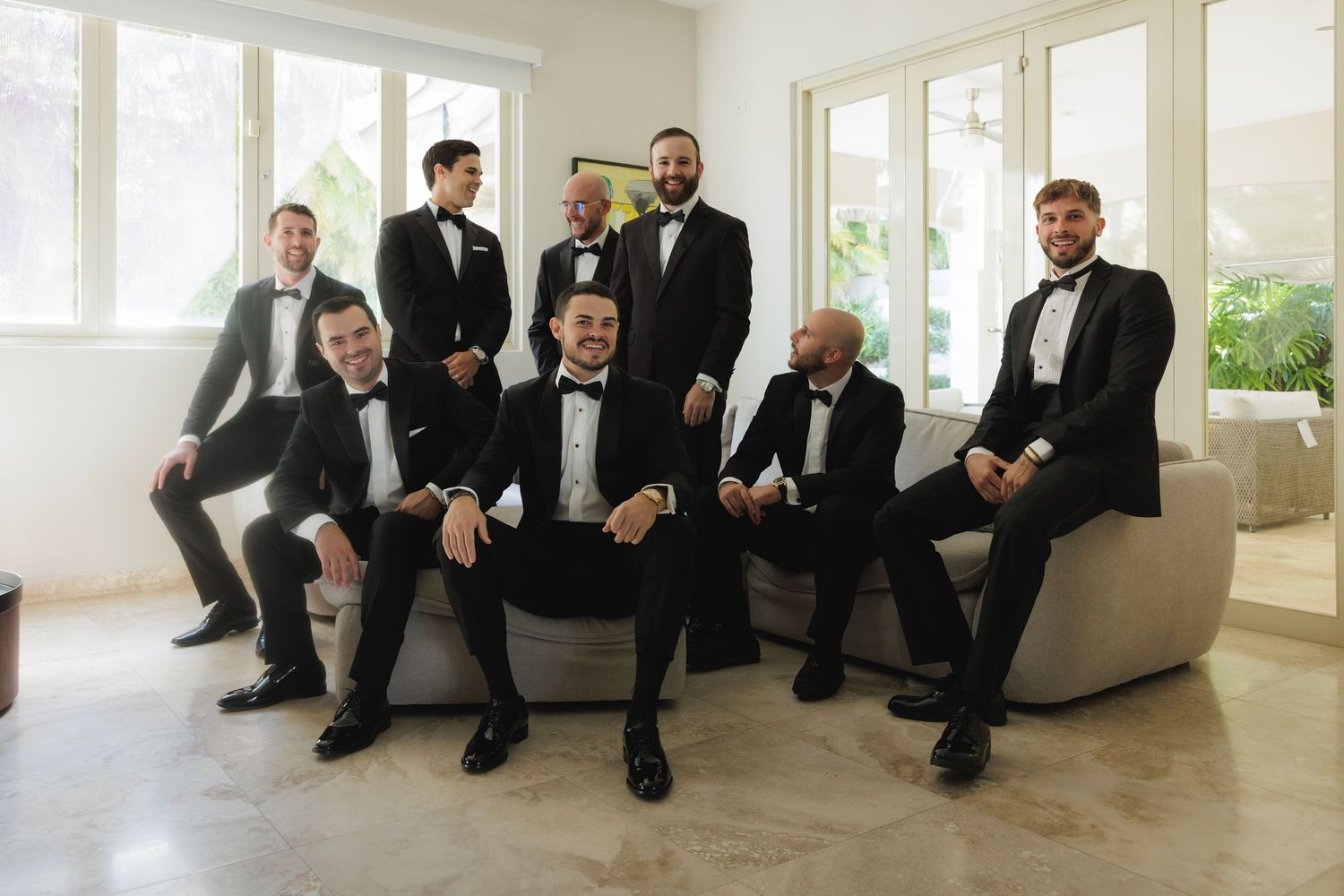Groomsmen in tuxedos relaxing on white furniture before wedding ceremony.