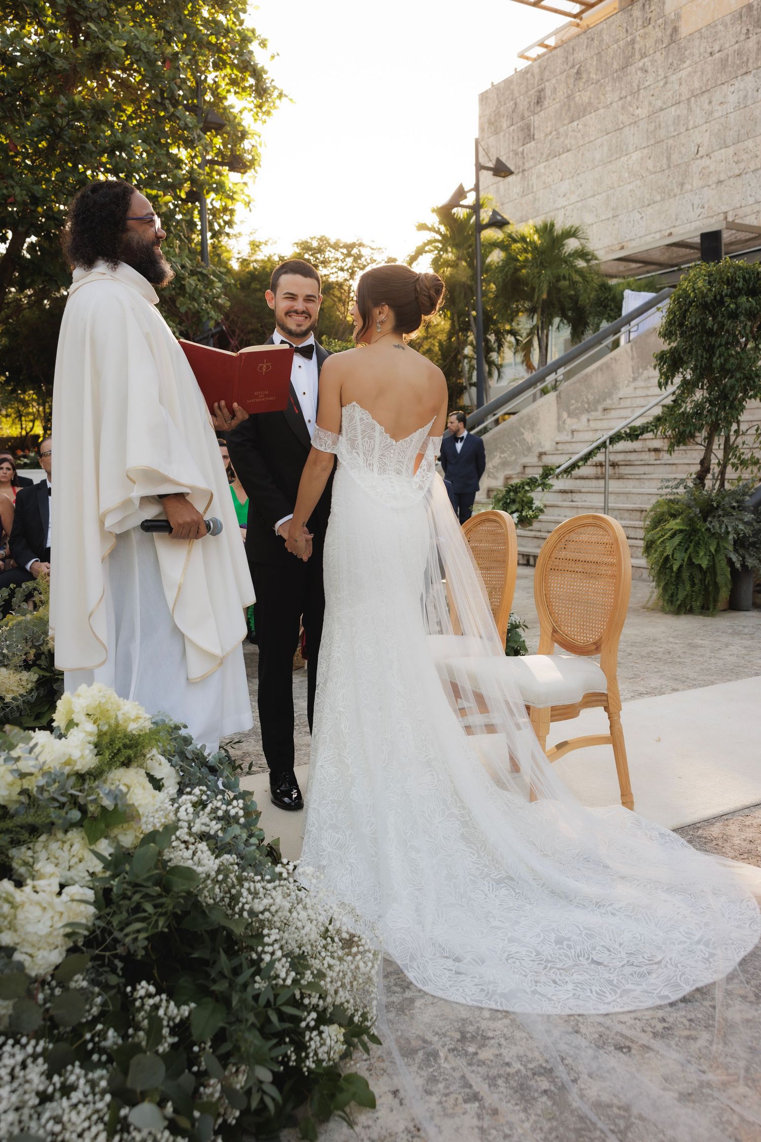 An intimate wedding ceremony moment with an officiant in white robes and ornate floral decorations.
