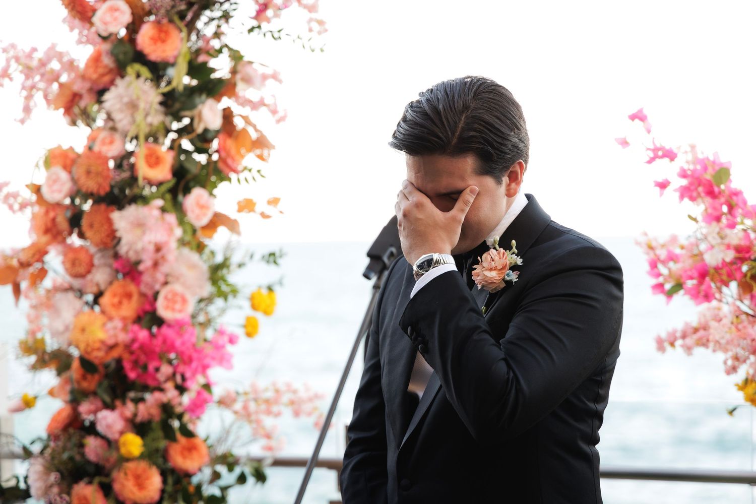 Emotional groom in black suit wipes eyes during wedding ceremony under floral arch with coral and pink blooms.