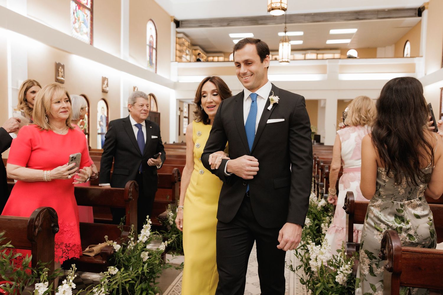 Wedding guests seated in church pews smile during an elegant ceremony with floral decorations along the aisle.