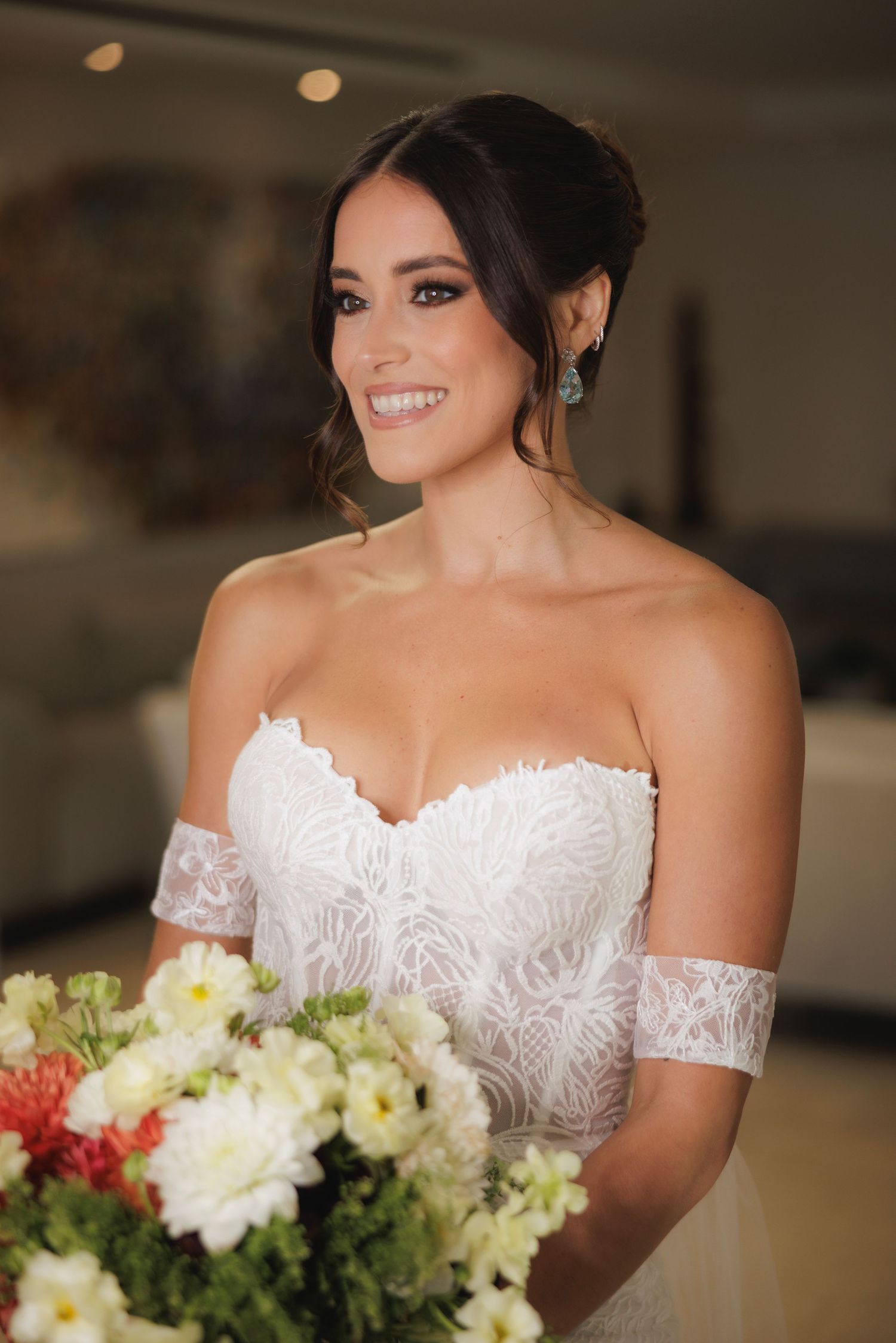 A bride wearing a white lace off-shoulder wedding gown smiles while holding a bouquet of white and pink flowers.