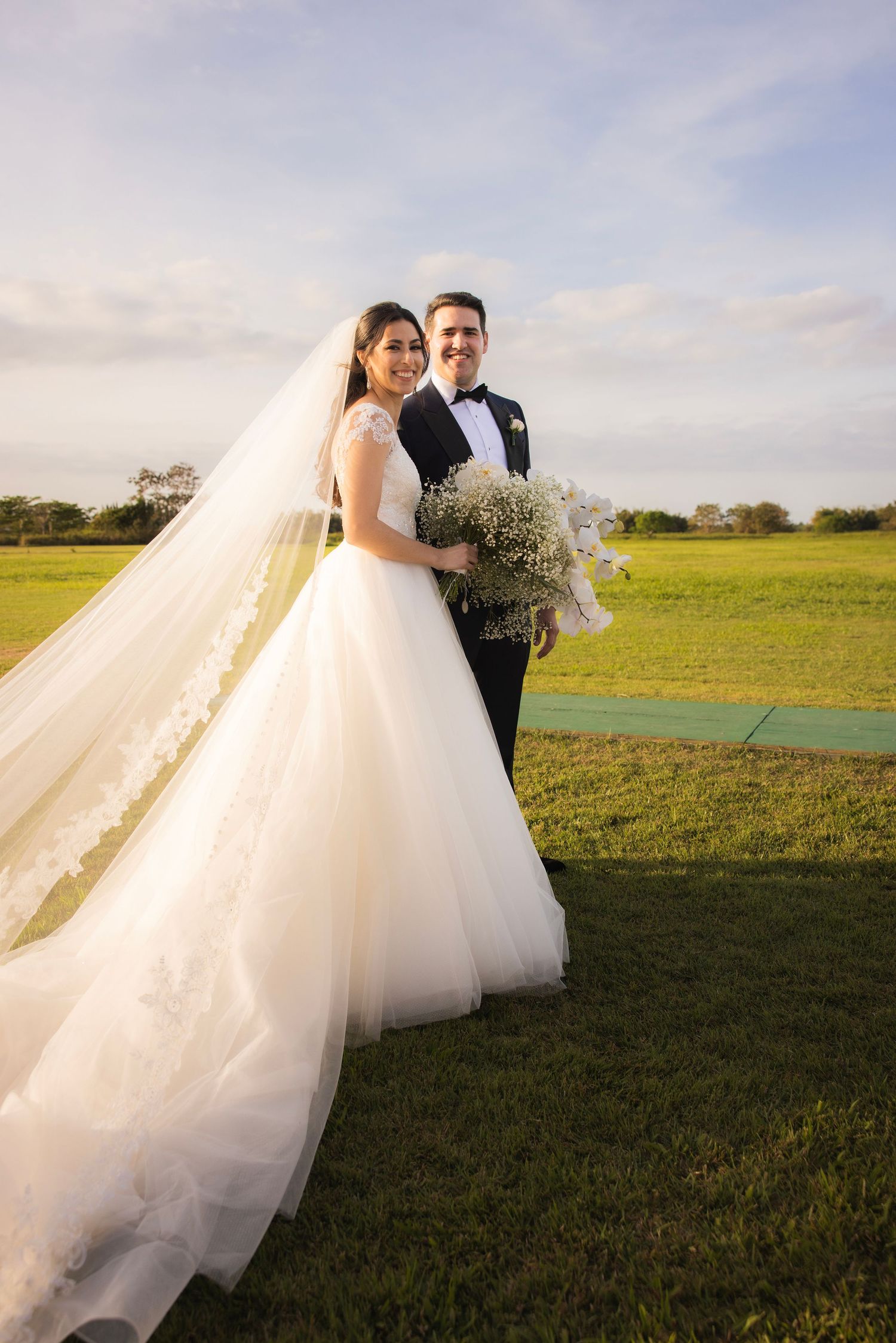 A beautiful wedding couple embraces in a sunny field with flowing white tulle dress trailing behind them during sunset.