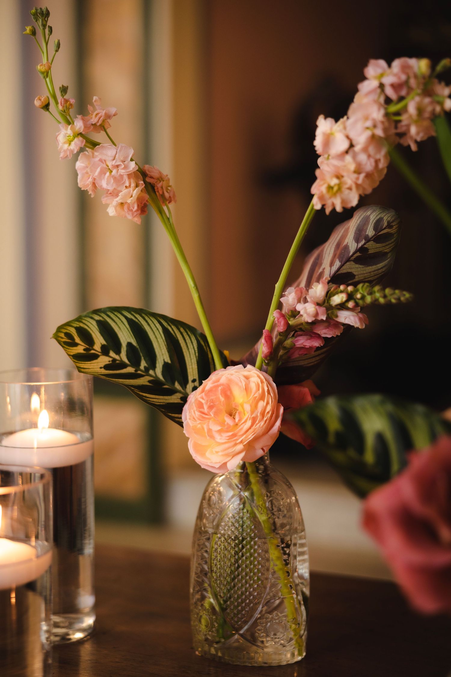 Glass vases with delicate pink flowers and candles create an intimate table setting with warm lighting and floral arrangements.