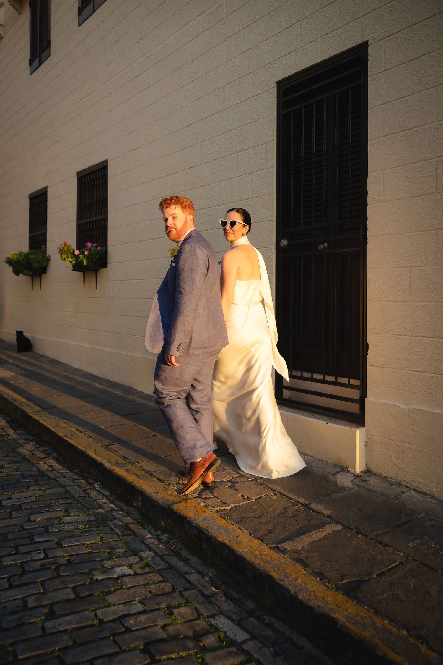 A couple walking along a cobblestone street beside a white colonial building.
