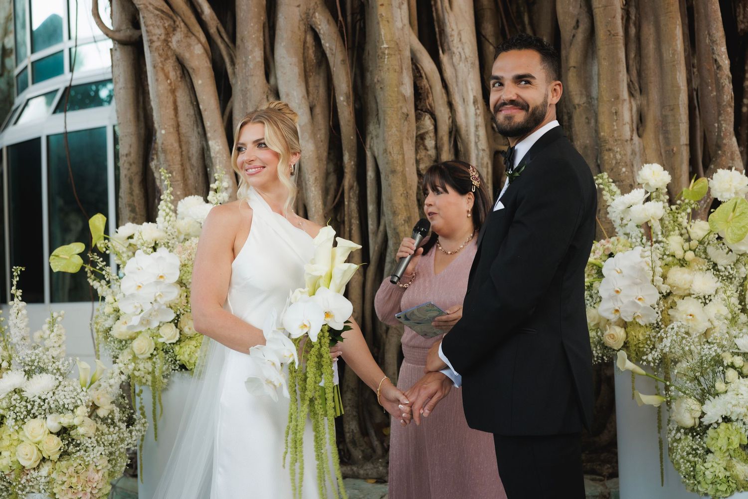 Outdoor wedding ceremony under a banyan tree with white floral decorations and a mauve-dressed officiant.