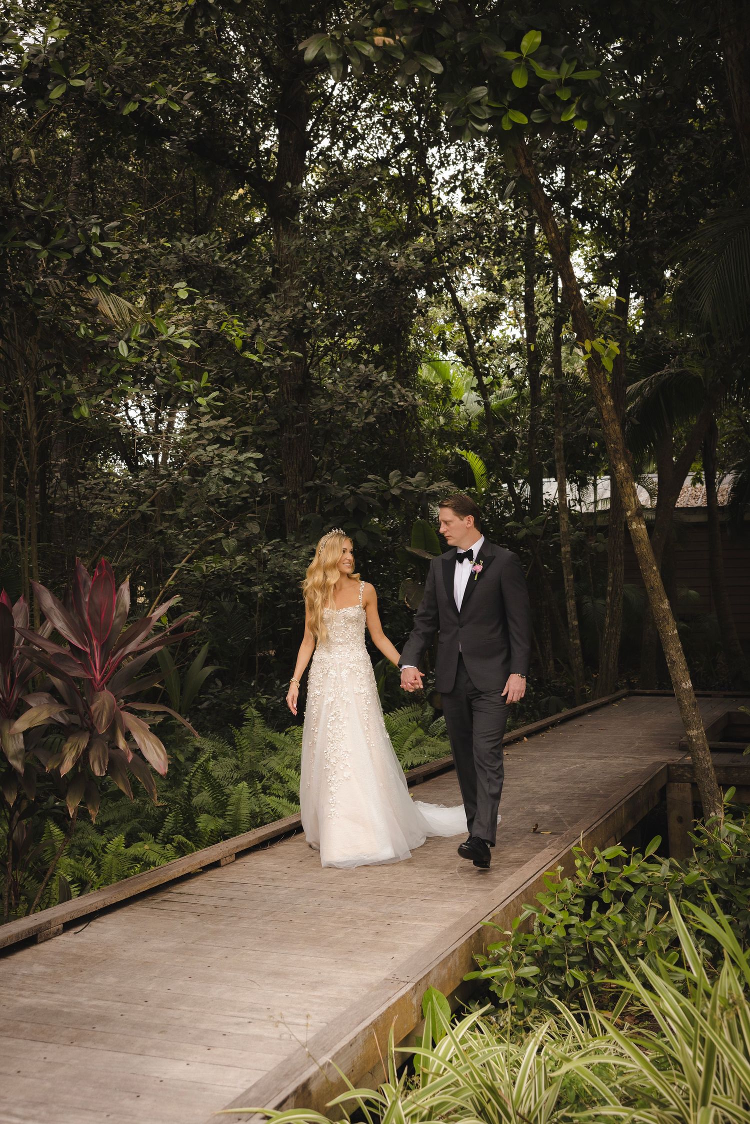 Wedding couple walking across a wooden bridge surrounded by lush tropical foliage in a garden setting at dusk.