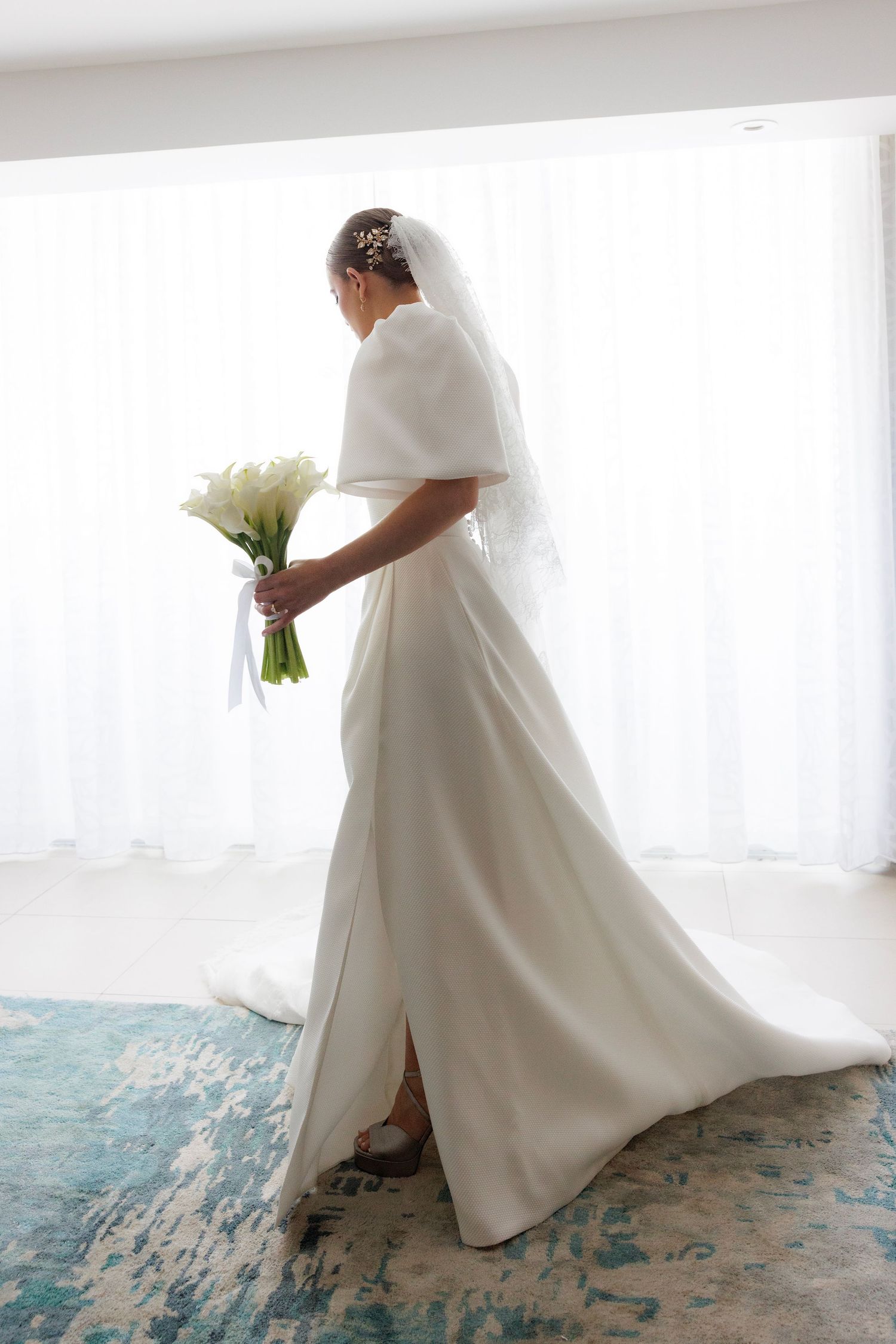 Elegant bride in flowing white silk dress with dramatic draped fabric poses against bright backlit window.