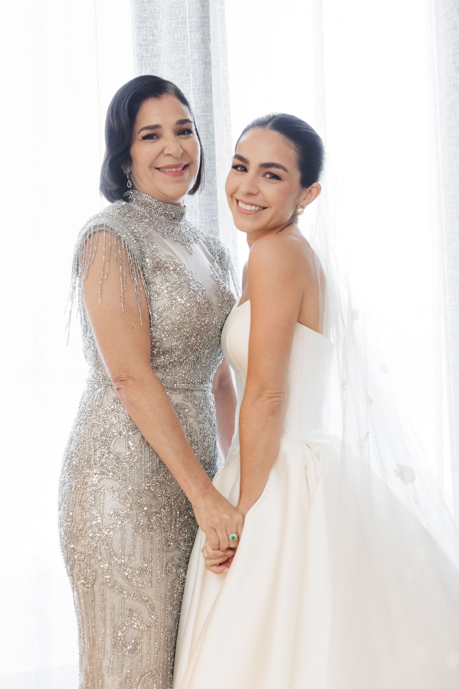 Two elegantly dressed women share a joyful moment together at a formal event by a bright window.