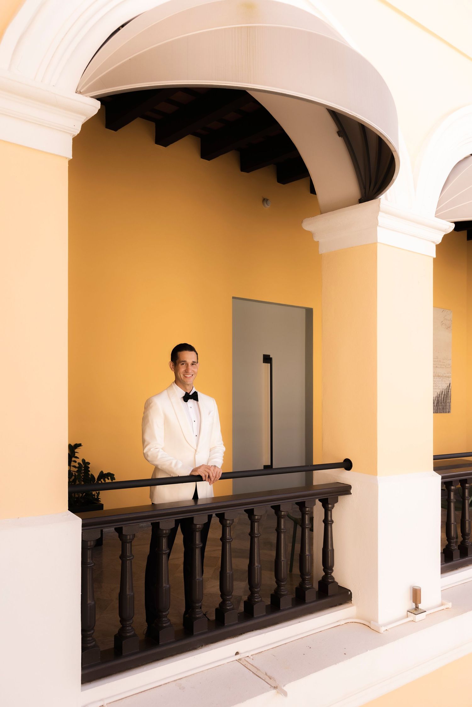 Elegant figure in a white gown standing in a classic colonnade walkway with yellow walls and dark railings.