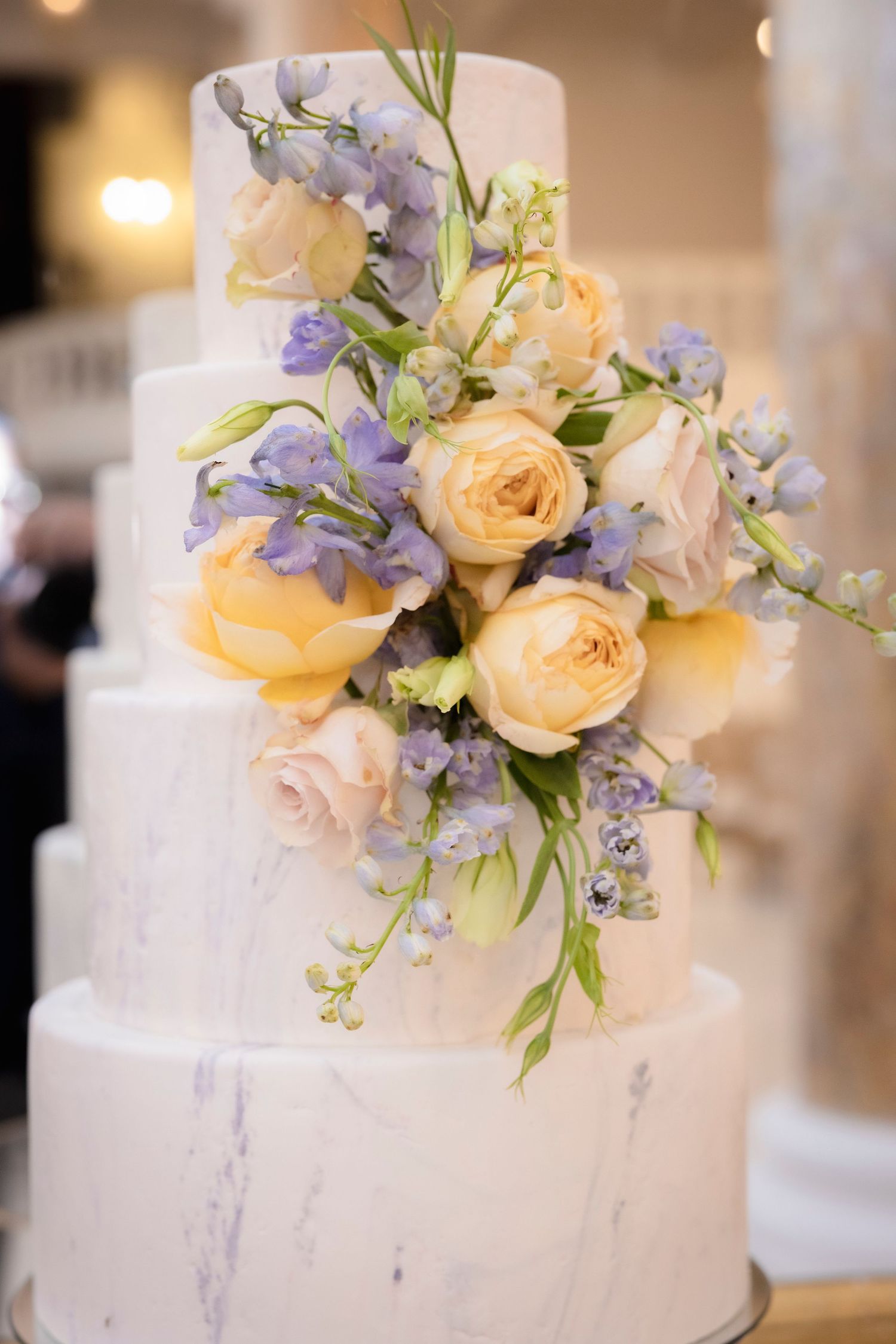 Elegant white wedding cake decorated with peach roses and purple delphinium flowers.