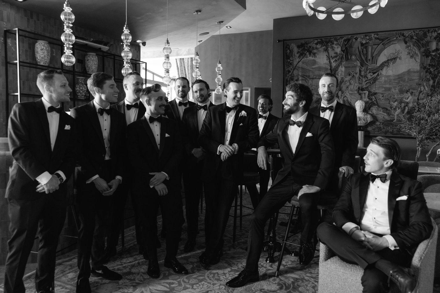A group in formal black tie attire stands together in a reception area with ornate decor and lighting fixtures.