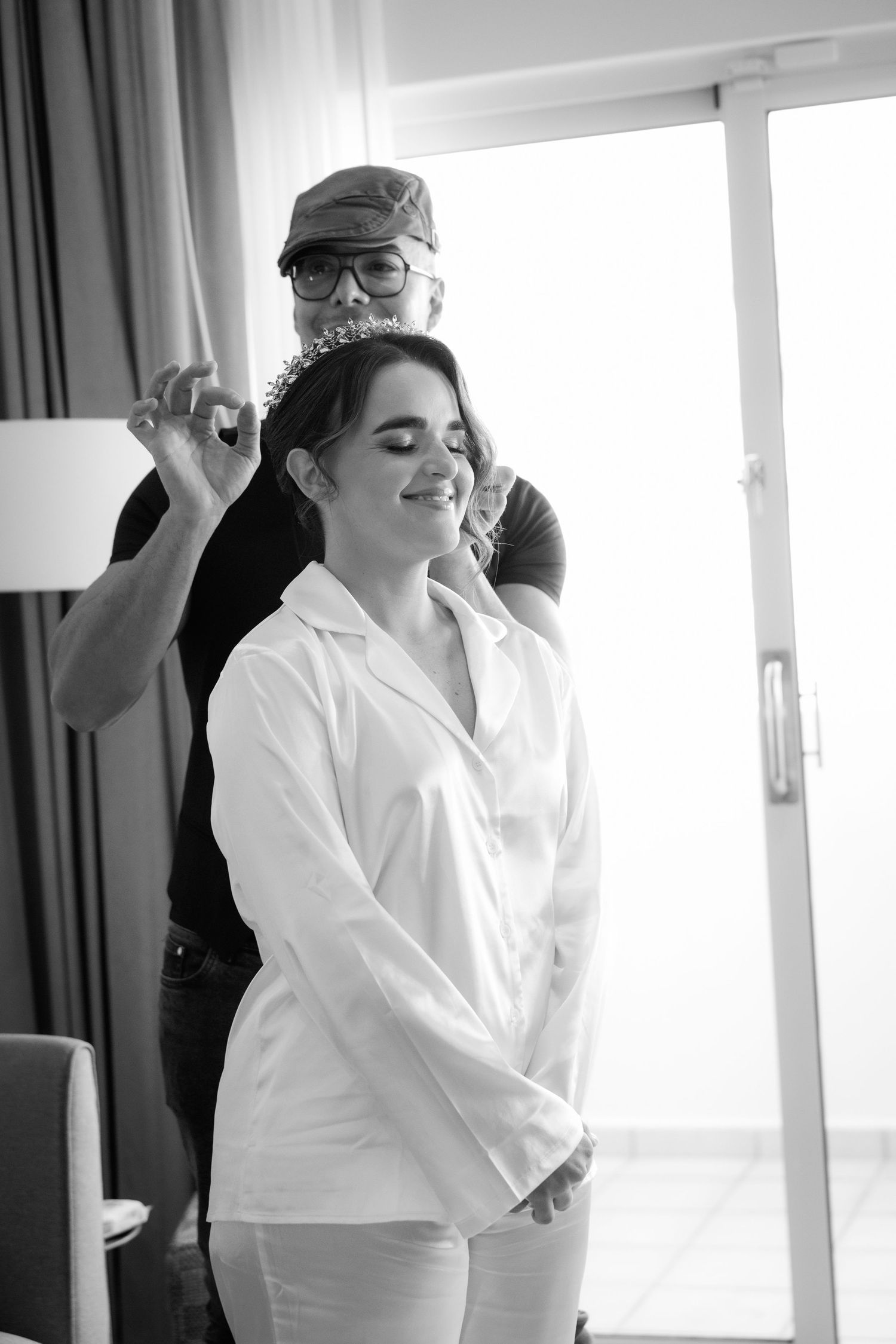 Wedding preparation scene showing someone getting their hair styled in a white robe by the window in black and white.