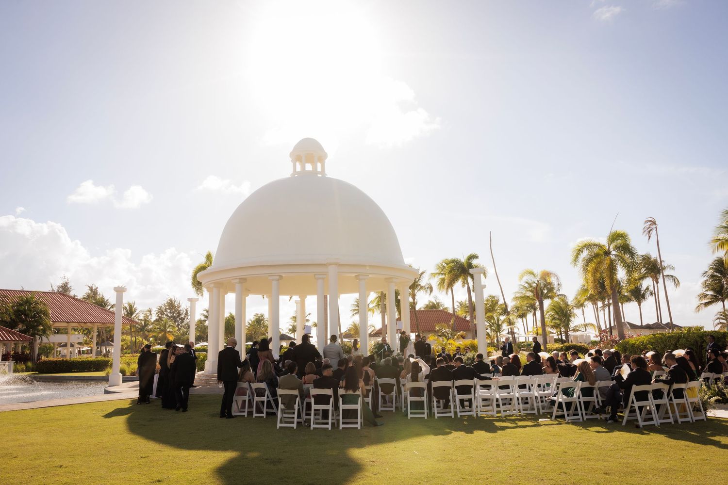 White gazebo wedding ceremony setup overlooking beach with palm trees and elegant seating on a sunny day.