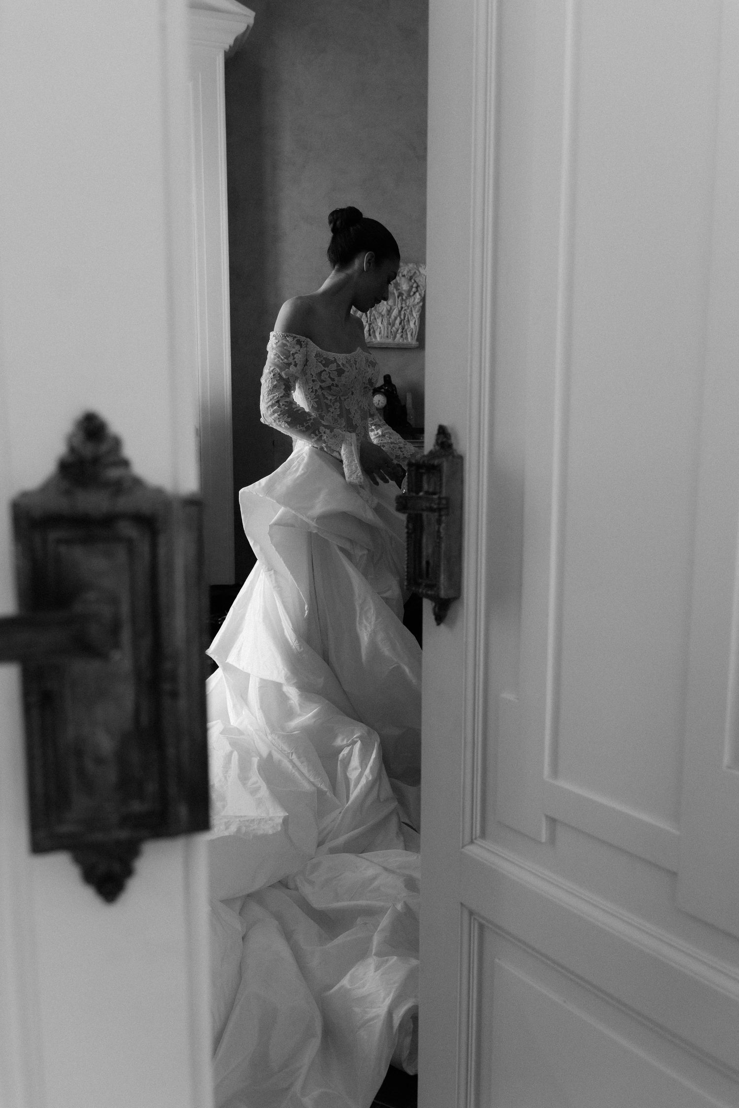 Black and white photo of a bride in a flowing white gown opening an ornate door, captured through a doorway.