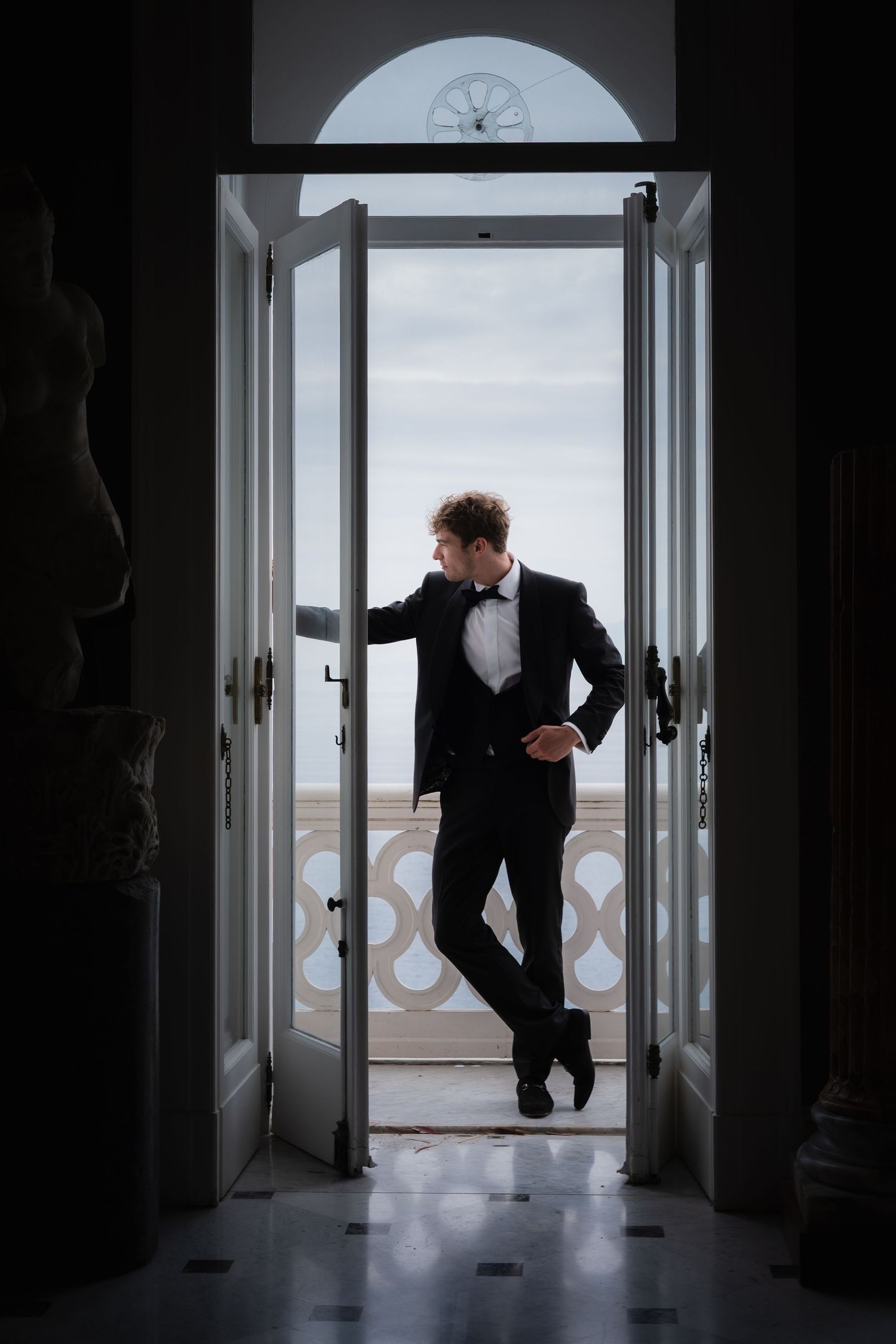 Elegant man in black tuxedo and bow tie leaning against open white doors overlooking a seaside balcony.