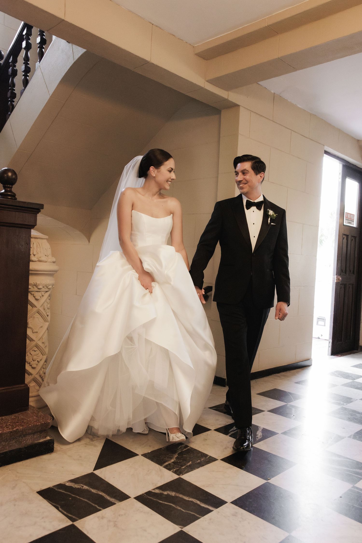 A couple in wedding attire walks together on a black and white checkered floor in an elegant hallway.