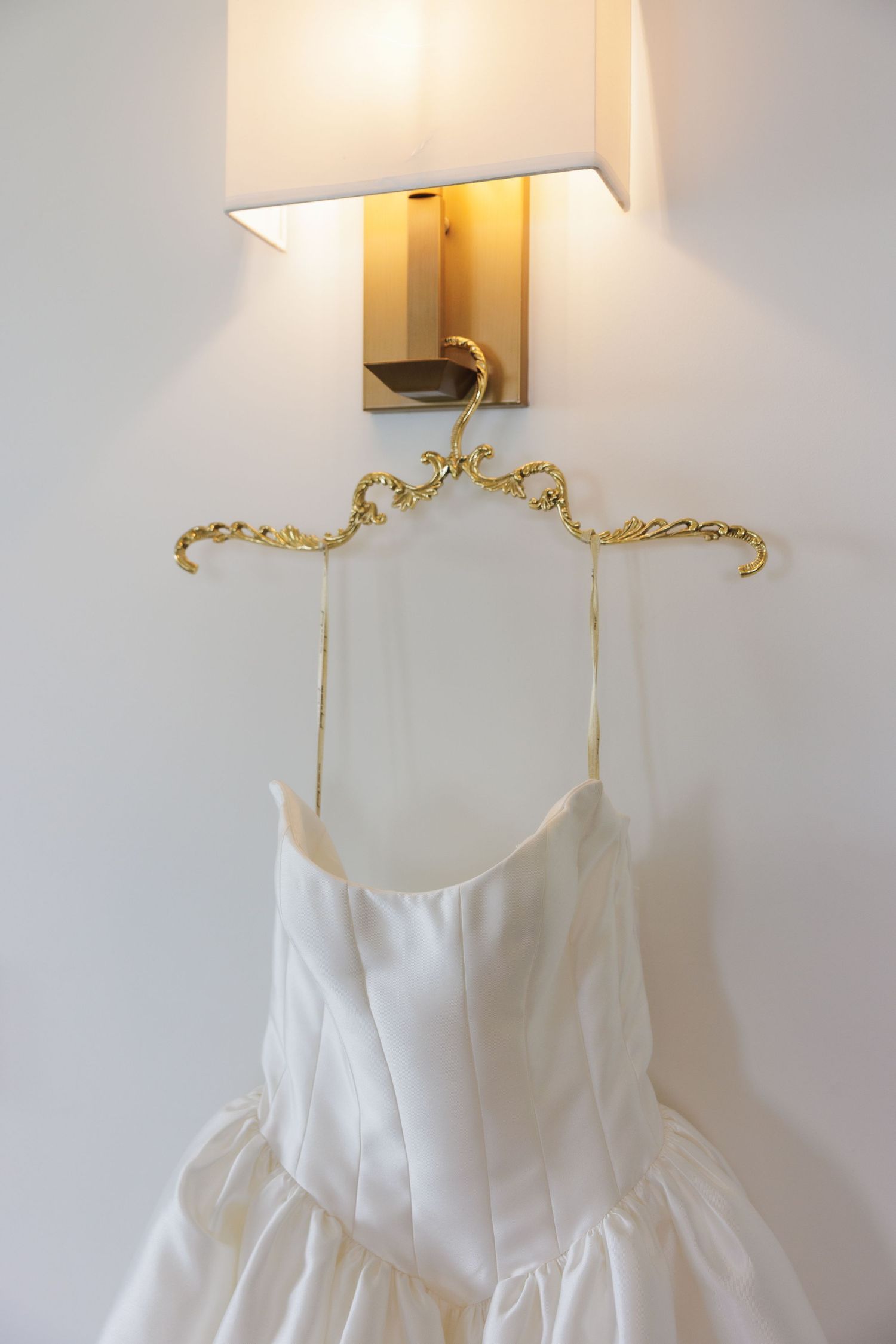 A white wedding dress hanging on an ornate gold hanger beneath a modern wall sconce.
