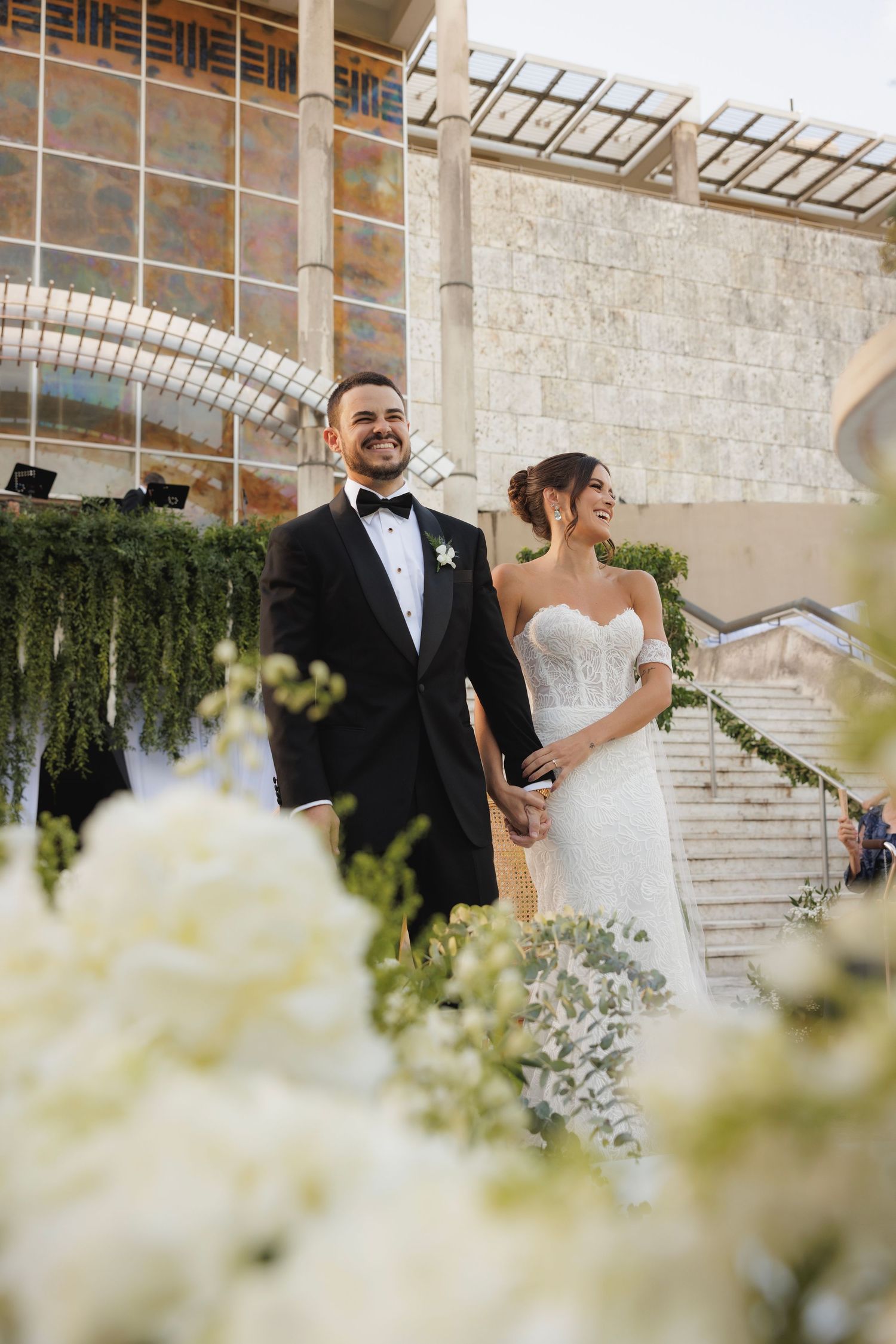 Newlyweds pose on grand stone staircase with white floral arrangements in foreground.