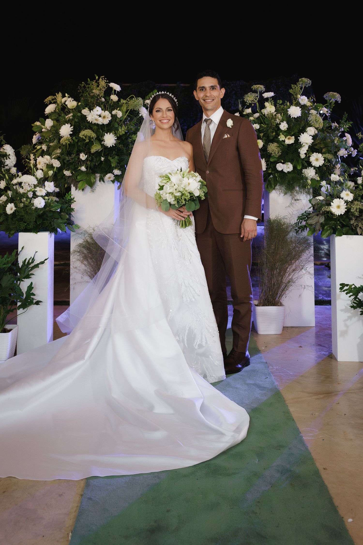 A wedding couple poses under an elegant floral arch with white flowers and greenery against a dark background.