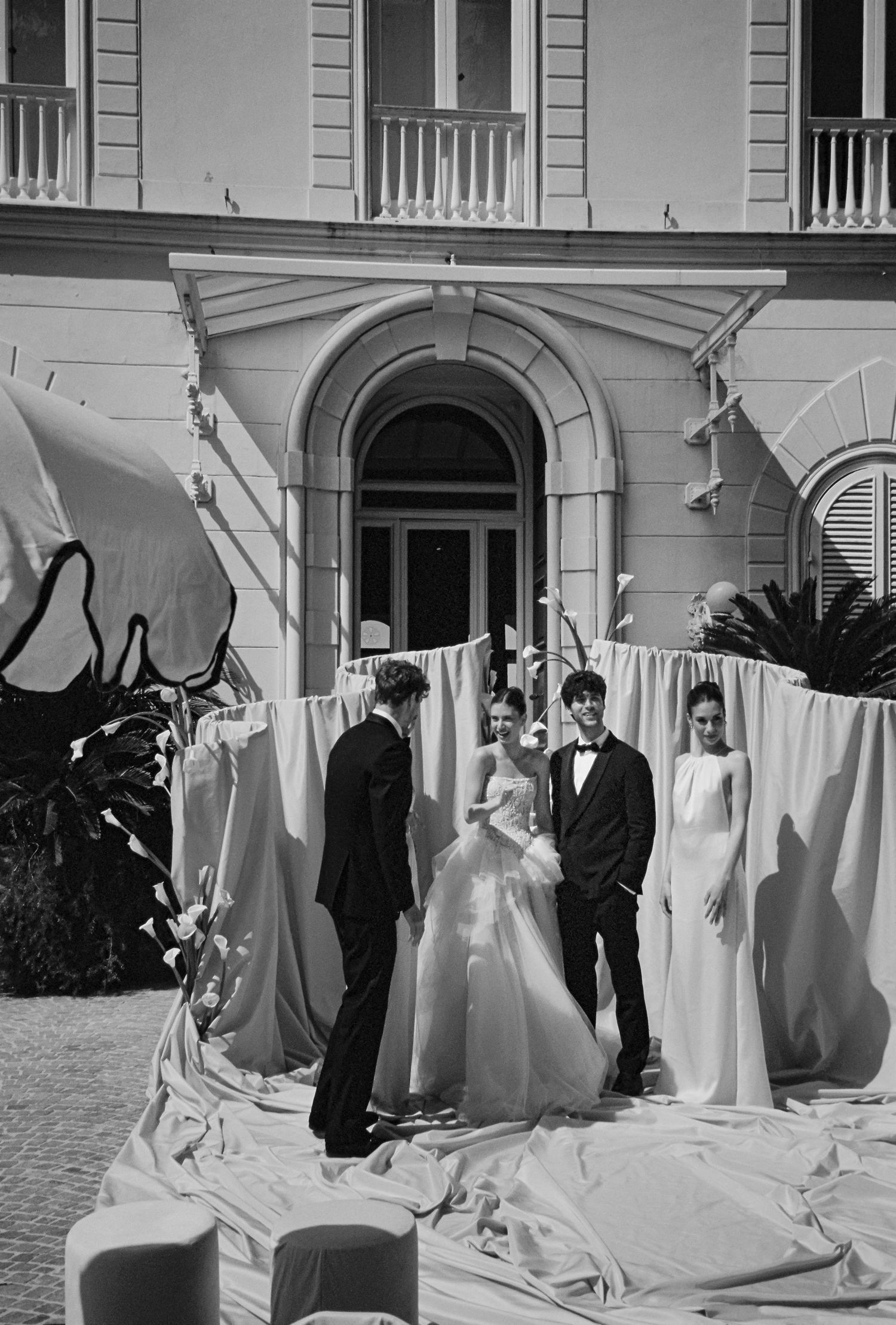 Black and white photo of wedding party in elegant gowns and tuxedos posing outdoors near draped fabric by a classic building.