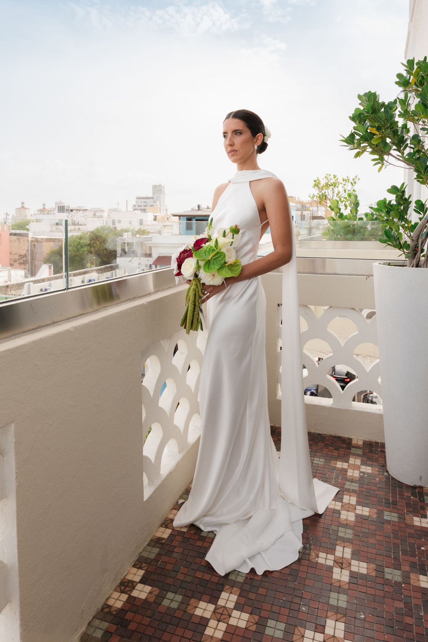 Elegant bride in white halter gown holding floral bouquet on rooftop balcony with city skyline backdrop.