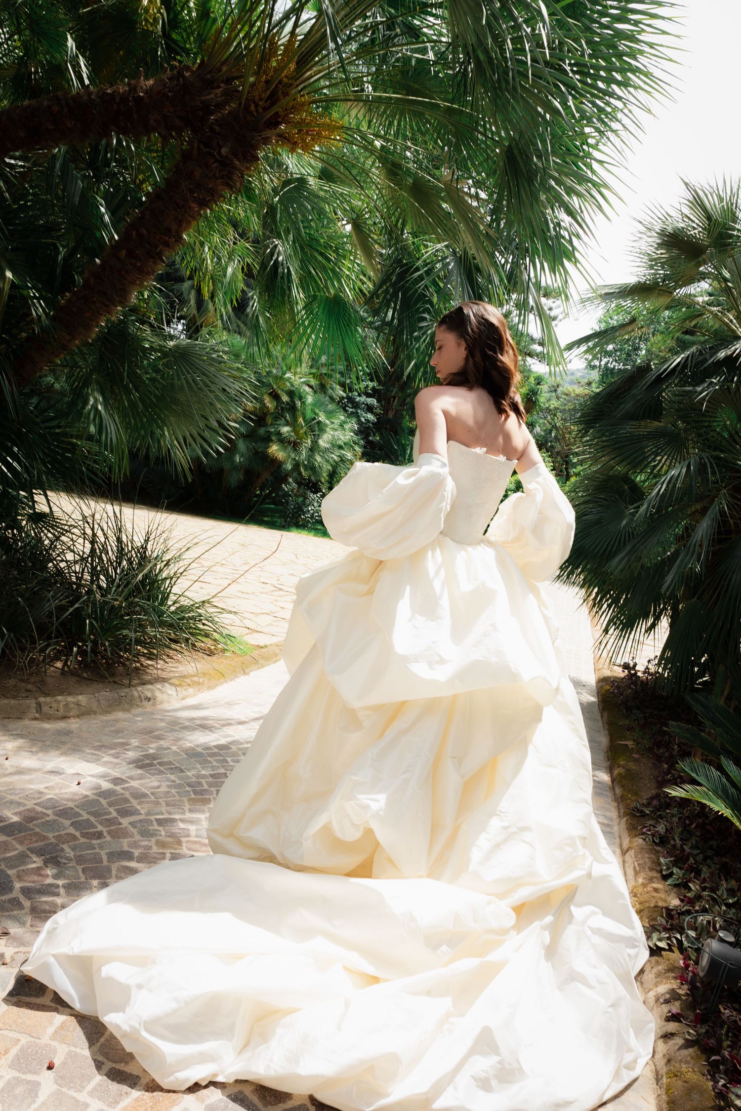Bride in voluminous ivory ball gown with puff sleeves walks along a sunlit garden path surrounded by lush palm trees.