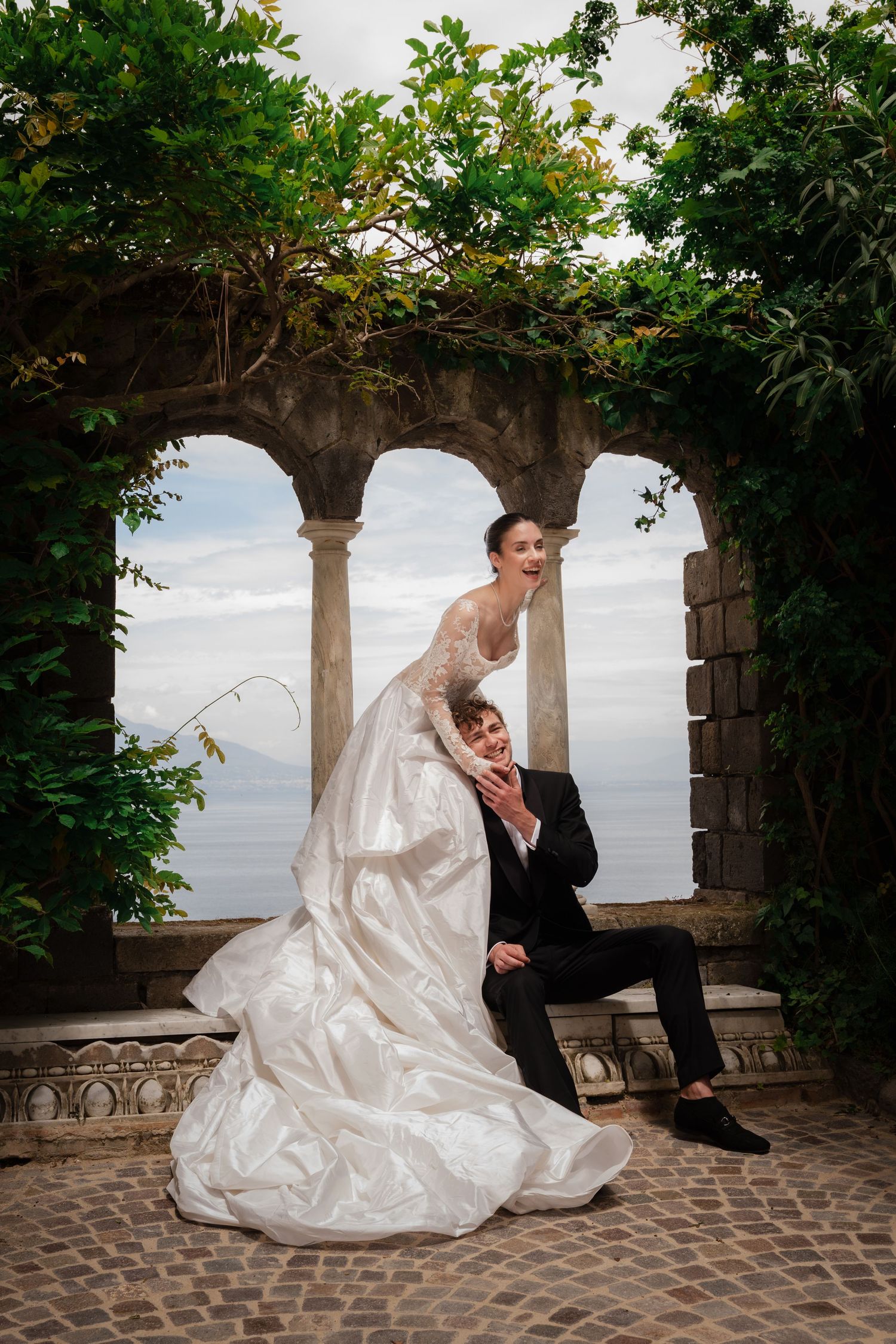 Joyful bride and groom posing under stone arches with lush greenery and ocean view at elegant wedding venue.