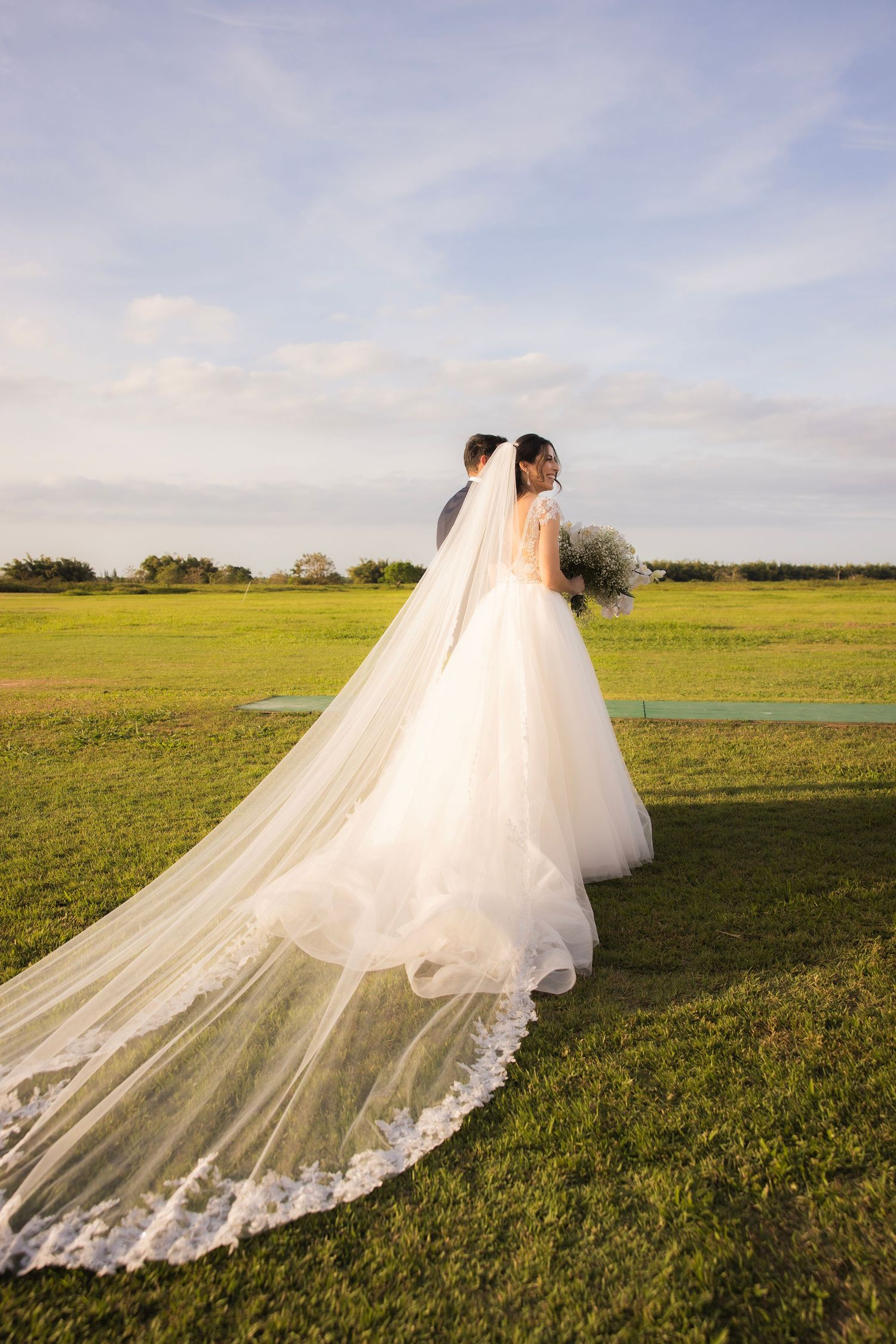 Flowing white wedding dress with lace veil trailing dramatically across a sunny green field.
