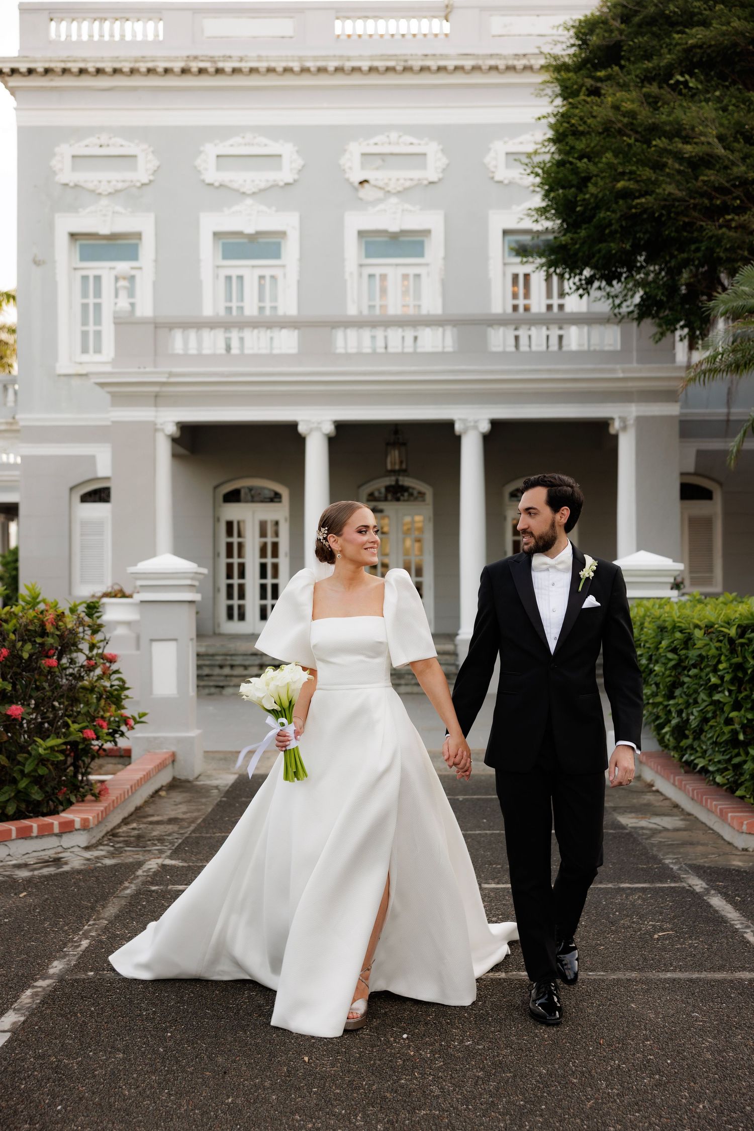 A couple in wedding attire walks together outside a grand white colonial mansion with ornate columns and architecture.