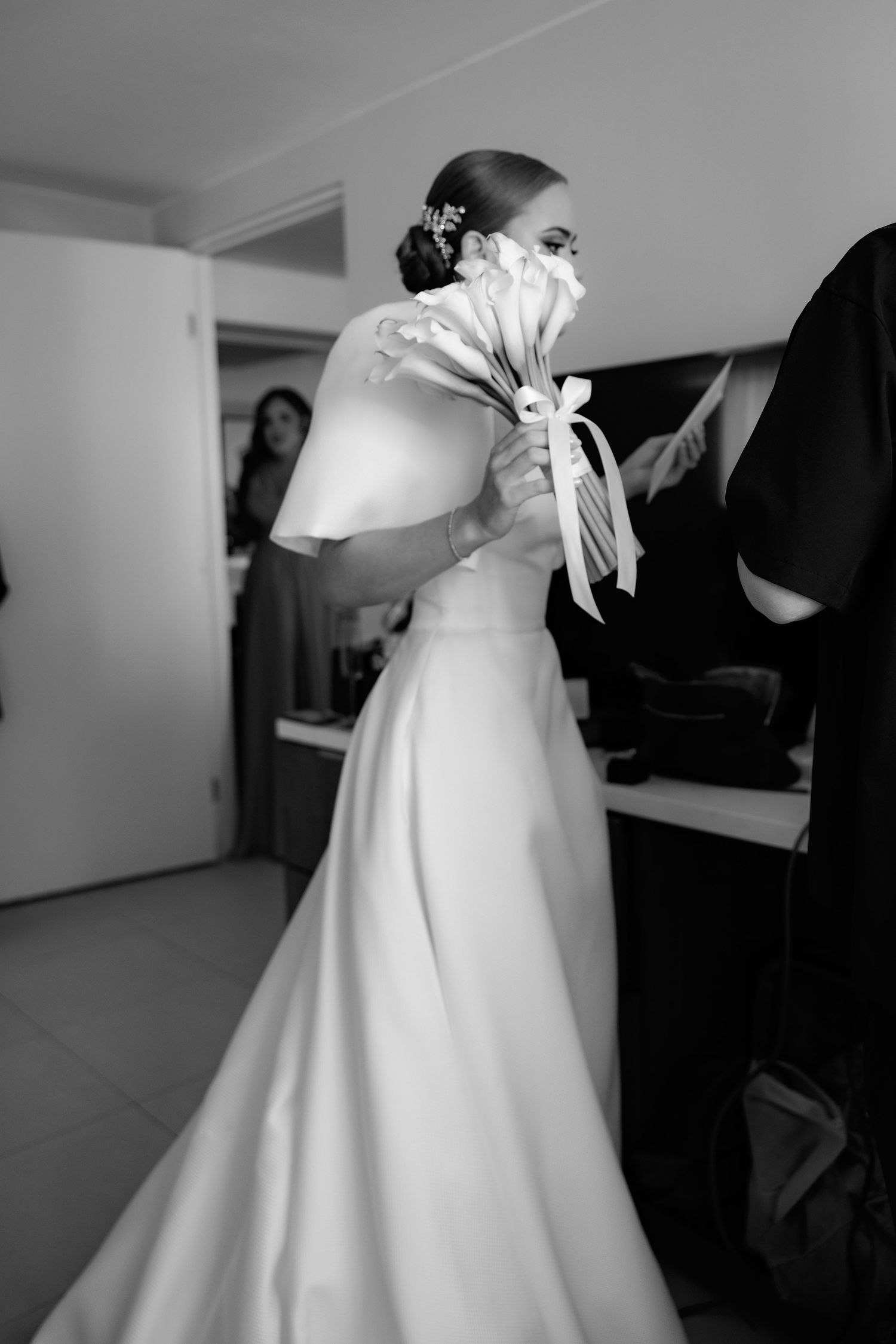 Black and white photo of a bride in an elegant white satin wedding gown with a bouquet during wedding preparations.
