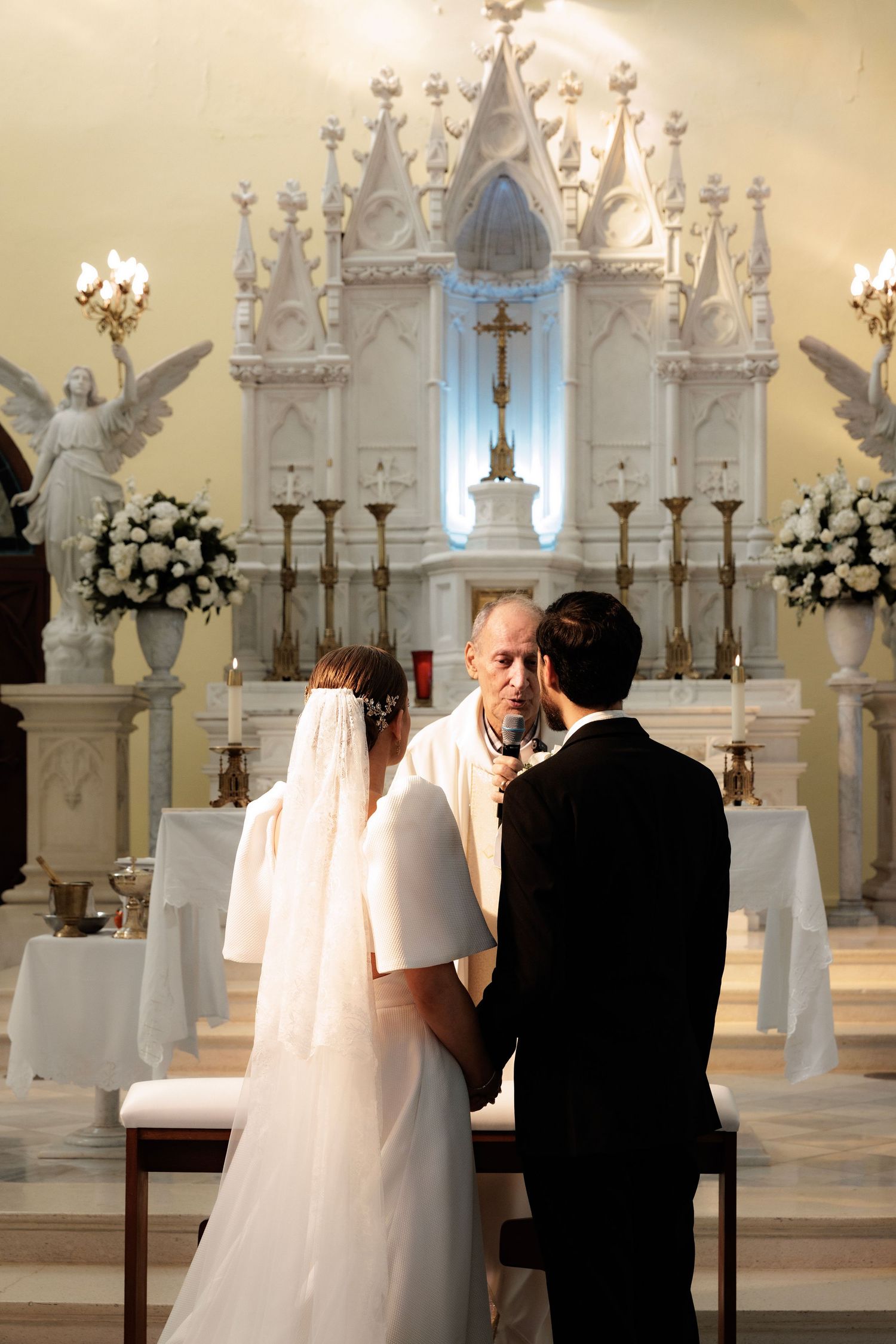 Wedding ceremony inside an elegant white cathedral with ornate Gothic architecture and multiple altar candles.