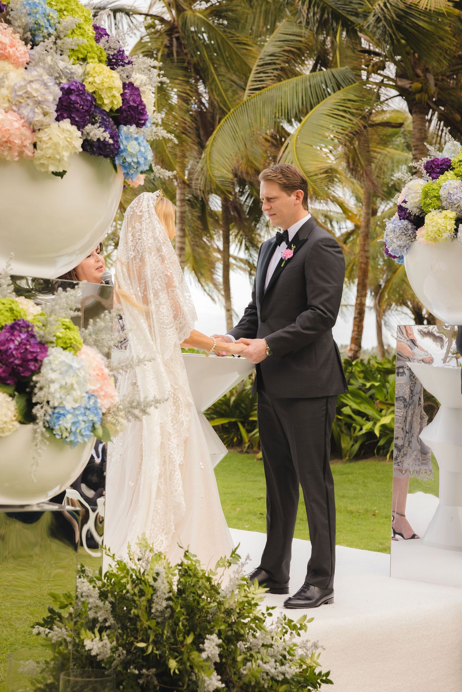 A tropical wedding ceremony under palm trees with elegant floral arrangements in white, purple and blue hydrangeas.