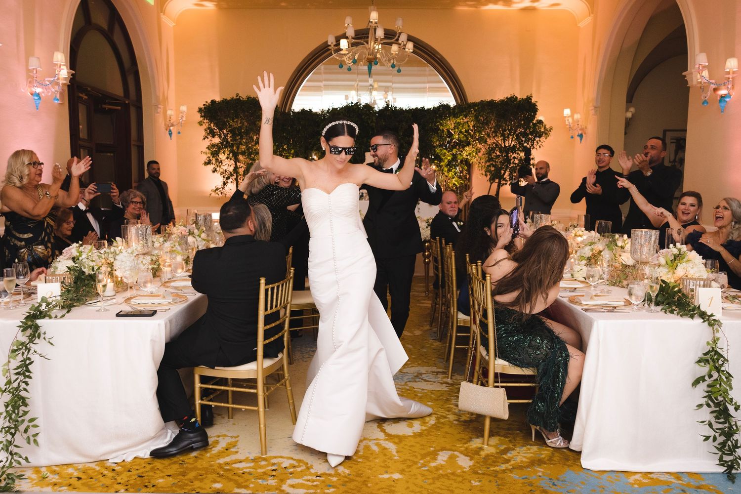 A wedding reception scene showing dancing in a grand ballroom with elegant table settings and floral decorations.
