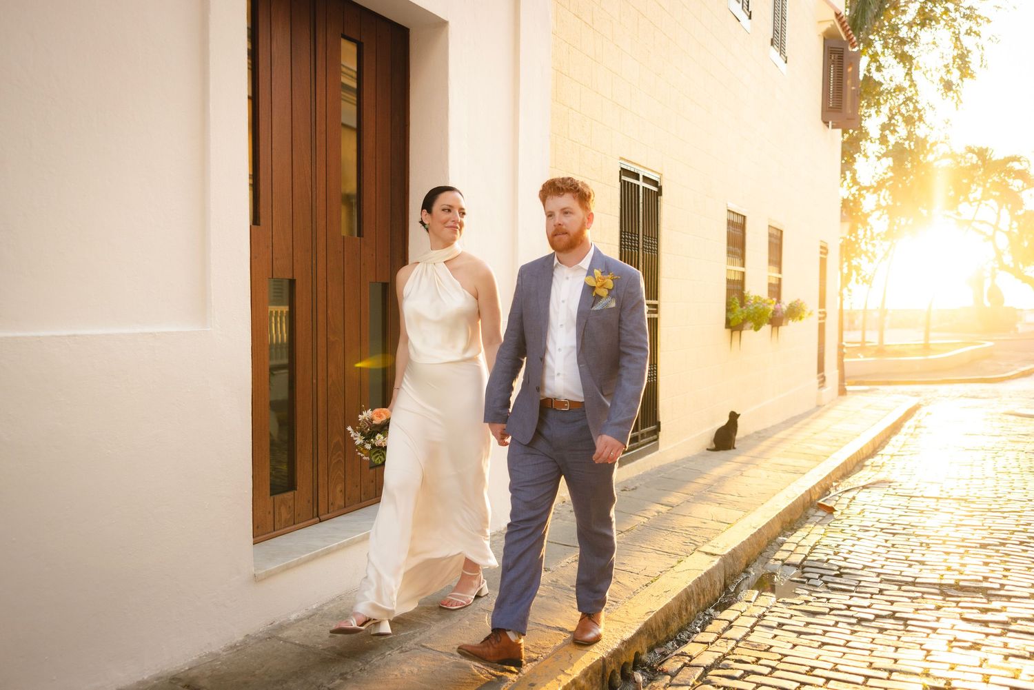 A couple walks down a sunlit cobblestone street in front of white Mediterranean buildings at sunset.