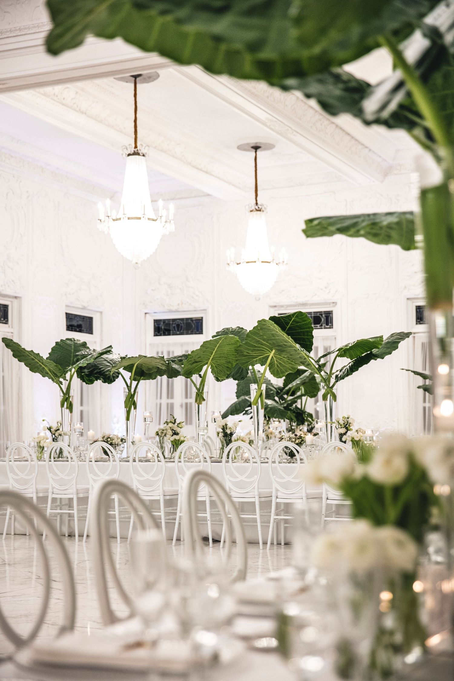 Modern white wedding reception venue with tropical palm leaves and elegant table settings in bright airy space.