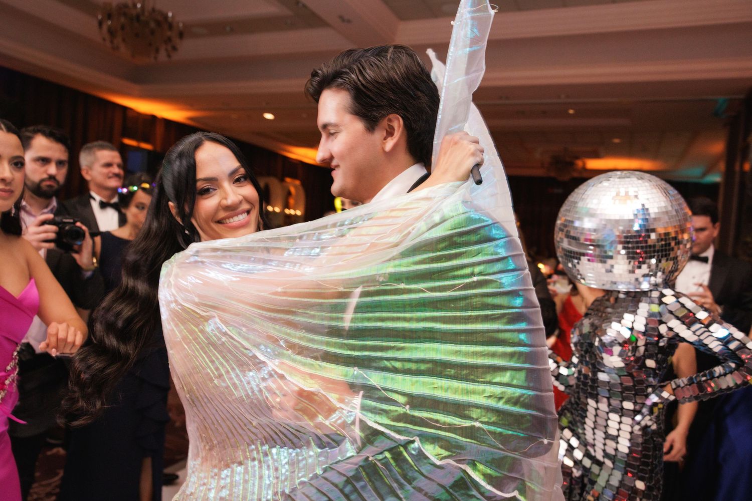 Wedding guests dance and celebrate together on the dance floor at a reception venue with a disco ball and warm lighting.