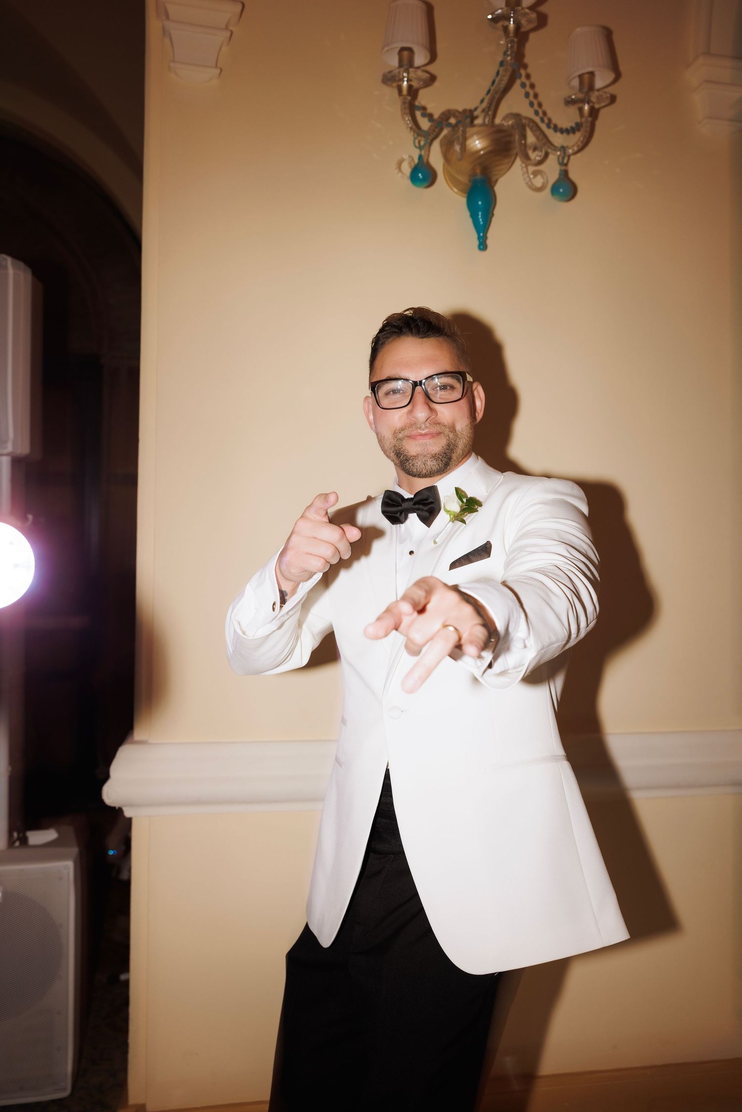 Person in white tuxedo jacket and bow tie making playful dance poses against beige wall.