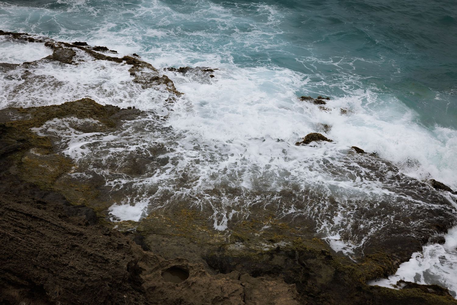 Aerial view of rugged coastline with turquoise ocean waves crashing against dark rocky cliffs.