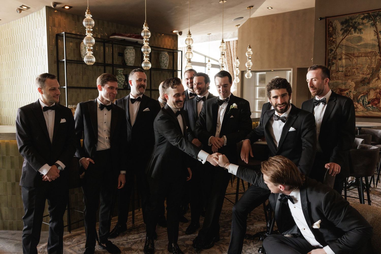 Groomsmen in black tuxedos raise a celebratory toast with whiskey glasses during a wedding reception.
