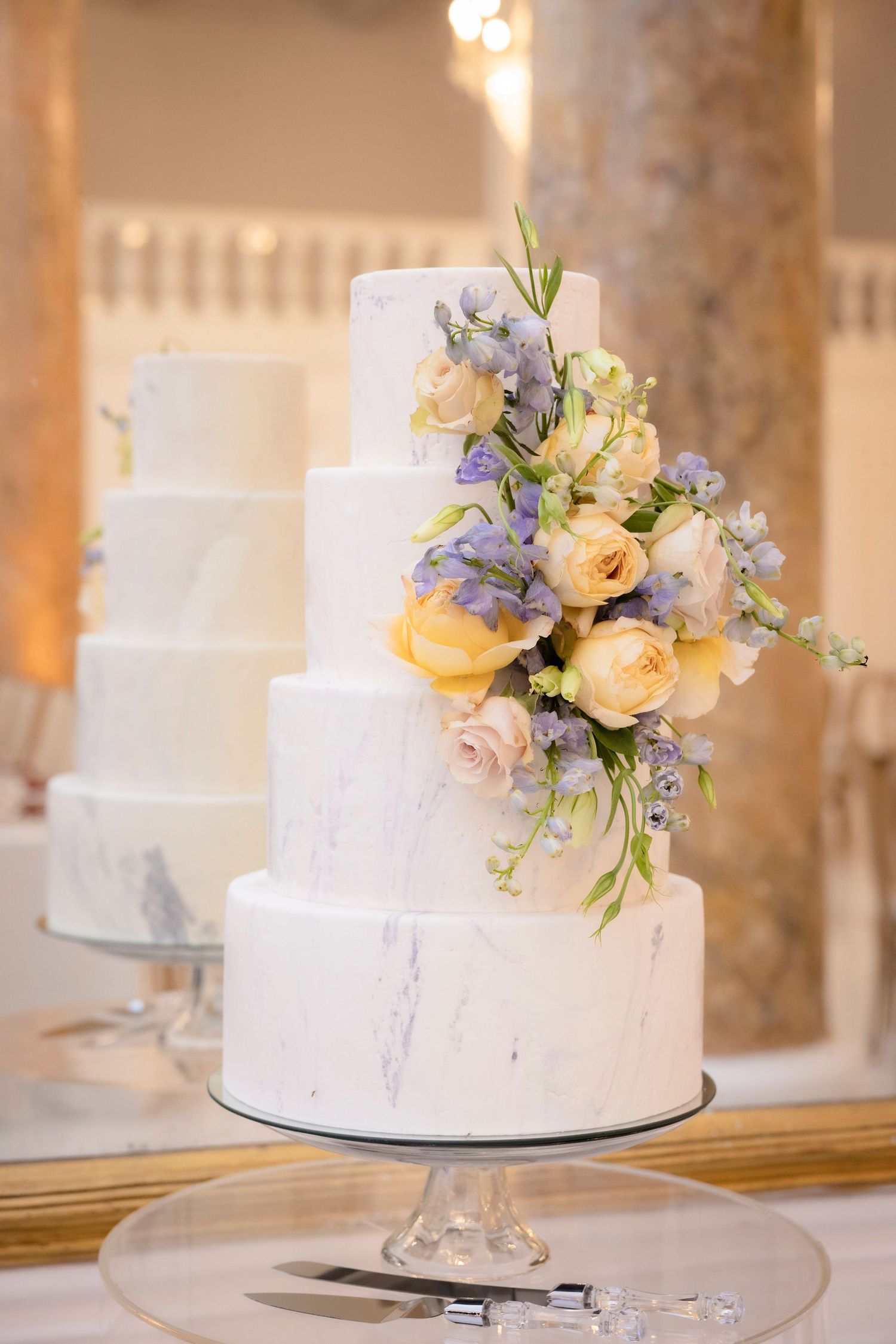 Elegant white marble-effect wedding cake with cascading peach roses and purple flowers on a crystal cake stand.