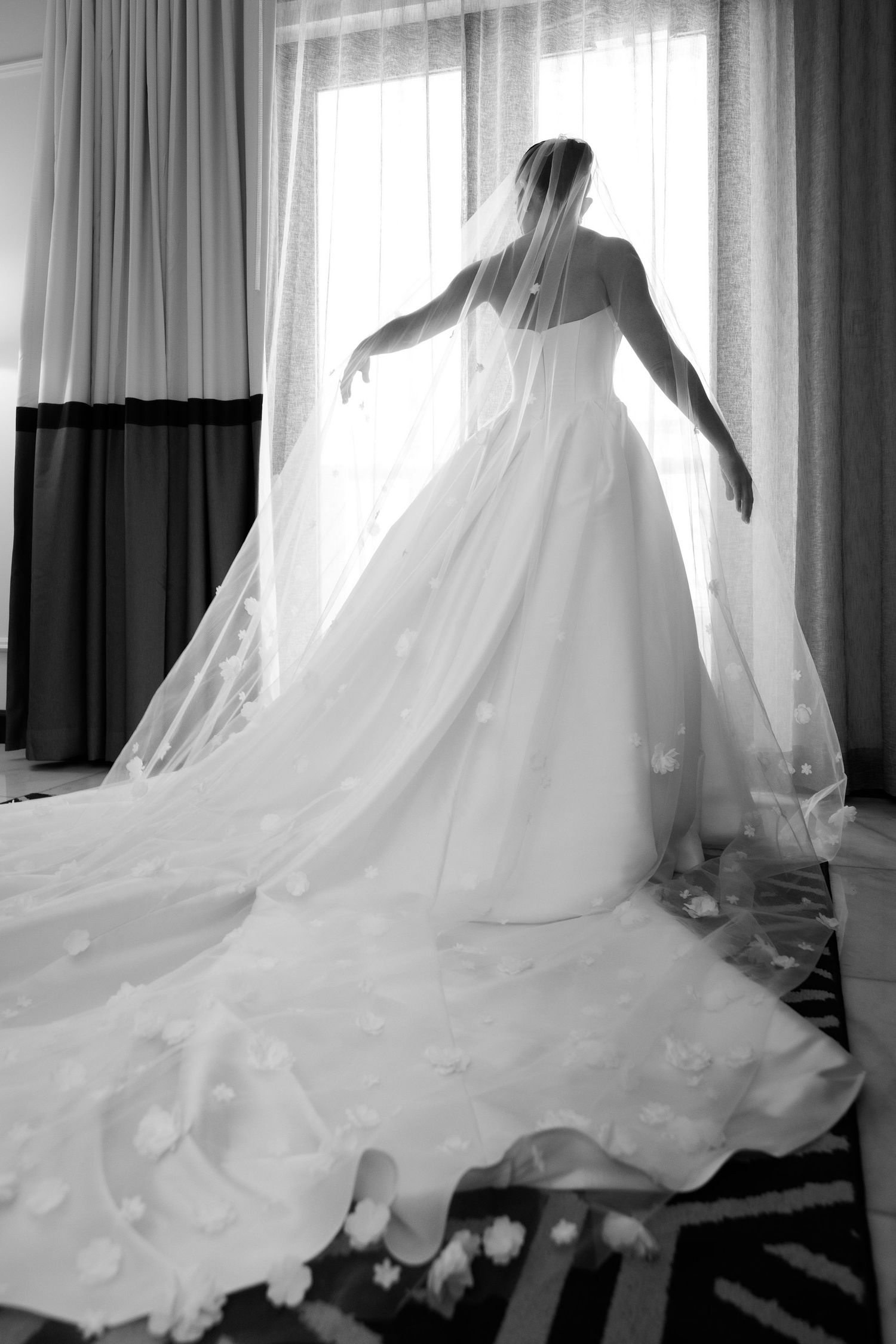 Silhouetted bride in flowing wedding gown and veil stands gracefully by window in dramatic black and white photo.
