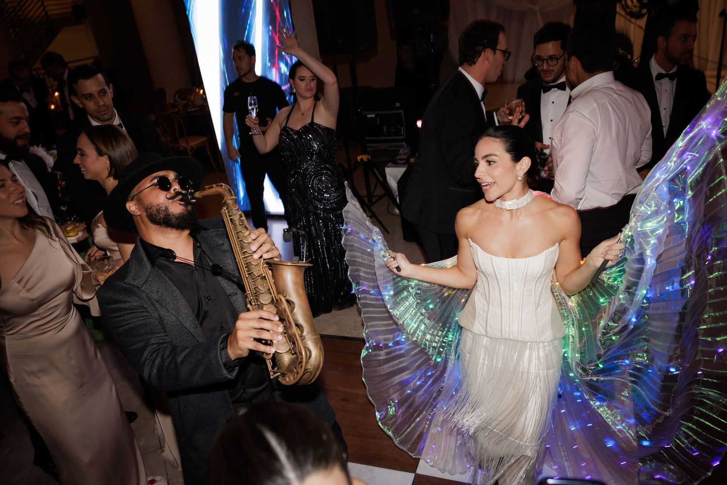 A saxophonist performs while a dancer with illuminated wings moves on the dance floor.