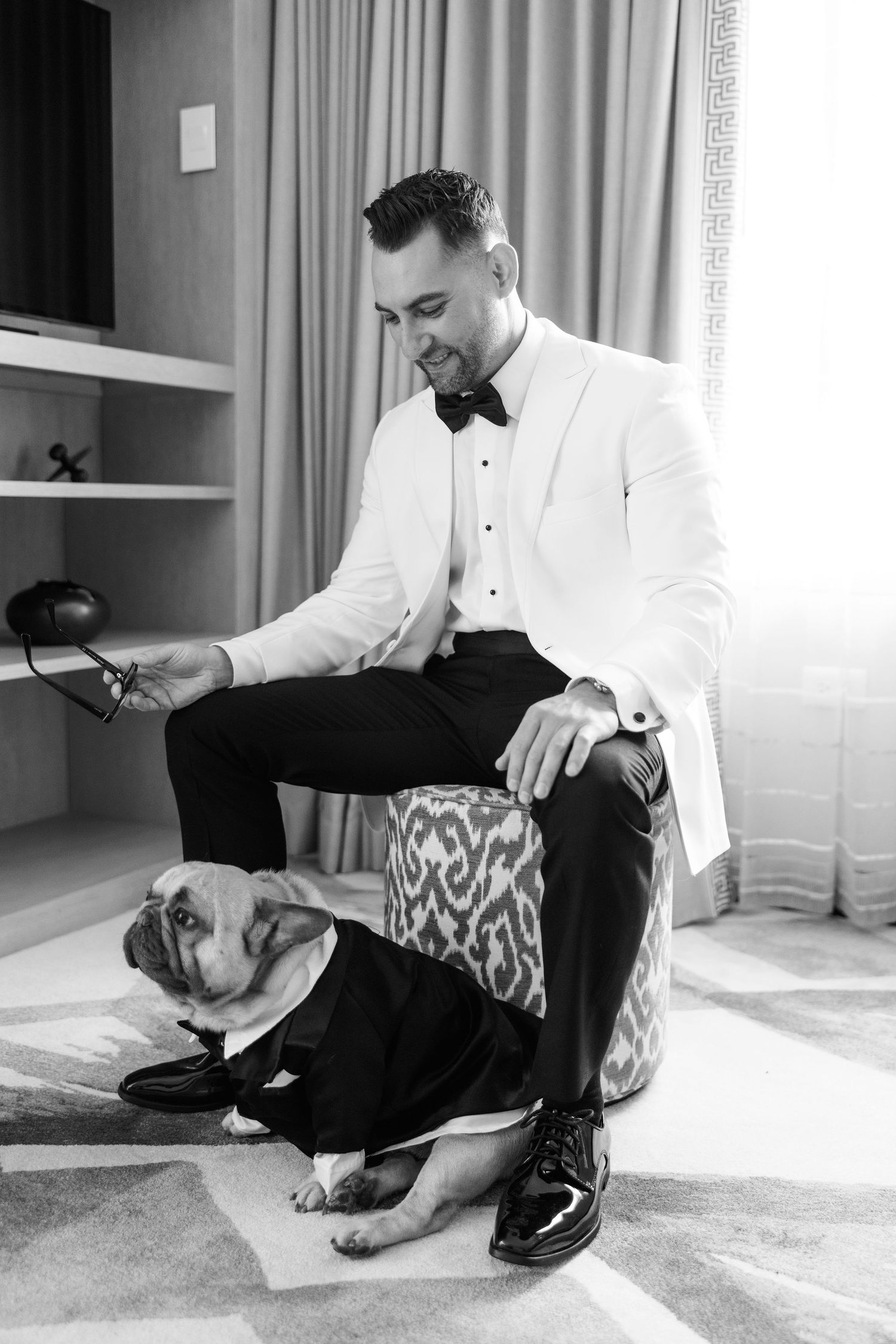 Two people in formal attire sharing a playful moment on the floor during wedding preparations.