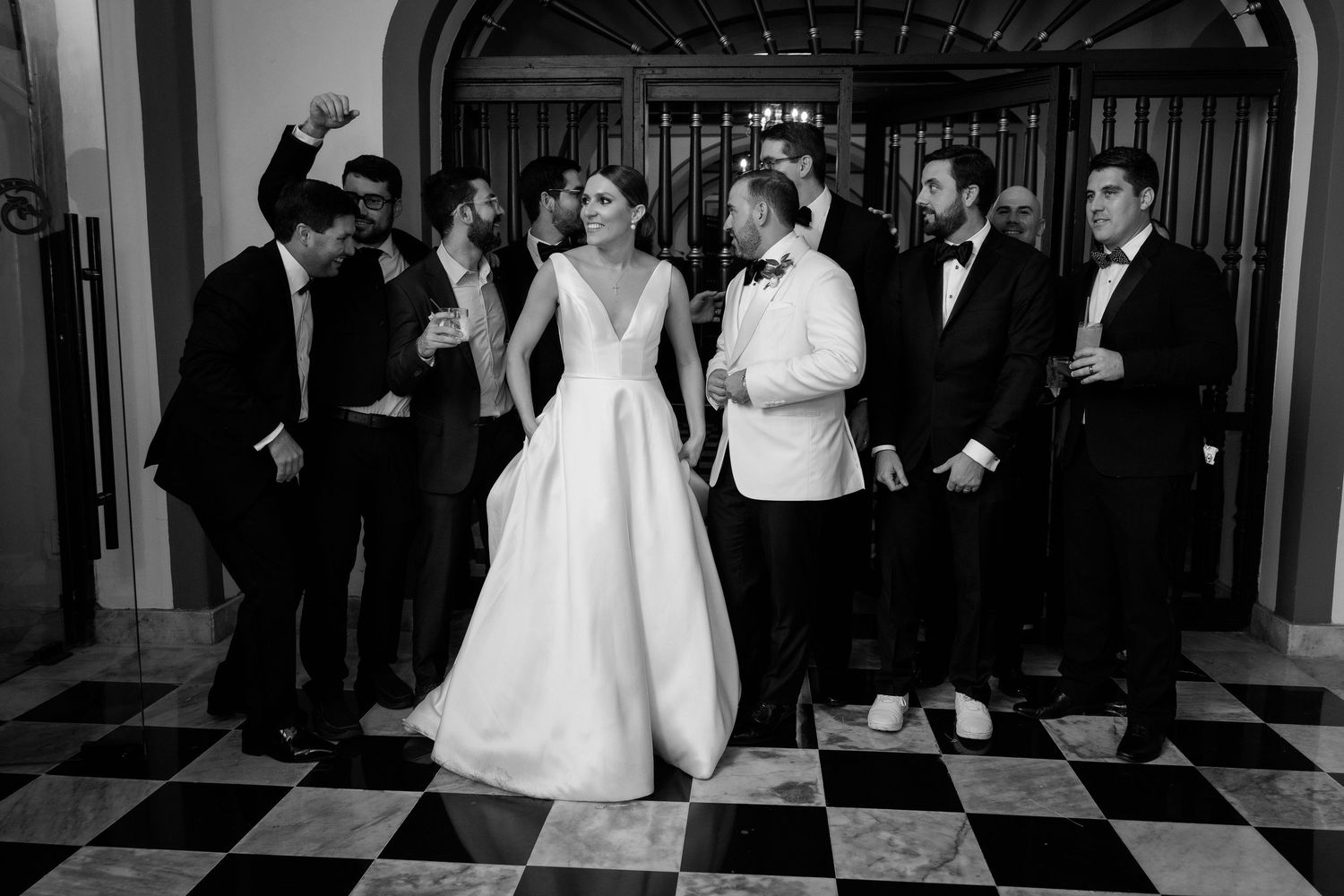 Wedding party in formal attire poses on black and white checkered floor at elegant venue.