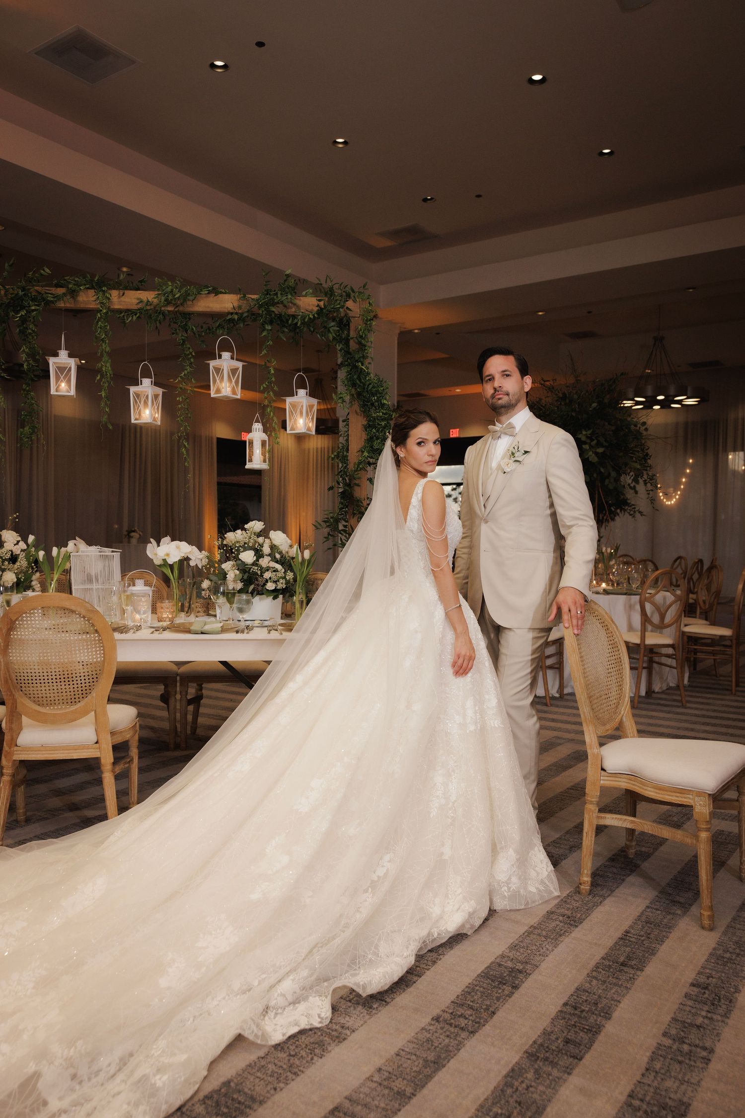 A couple in formal wedding attire stands elegantly in a luxurious ballroom with lantern decorations and plush seating.