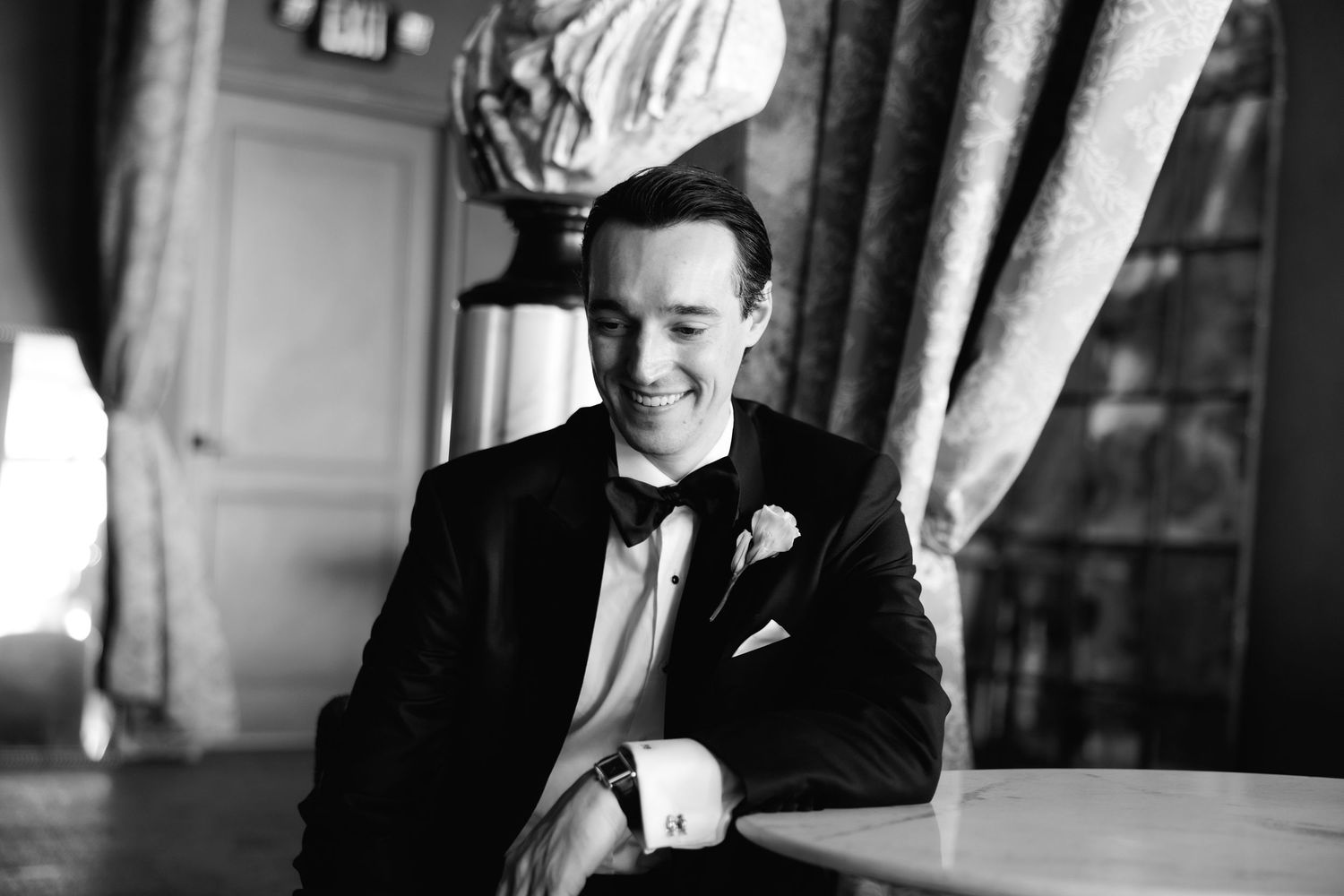 A groom in a classic black tuxedo and bow tie smiles warmly in an elegant indoor setting, captured in black and white.