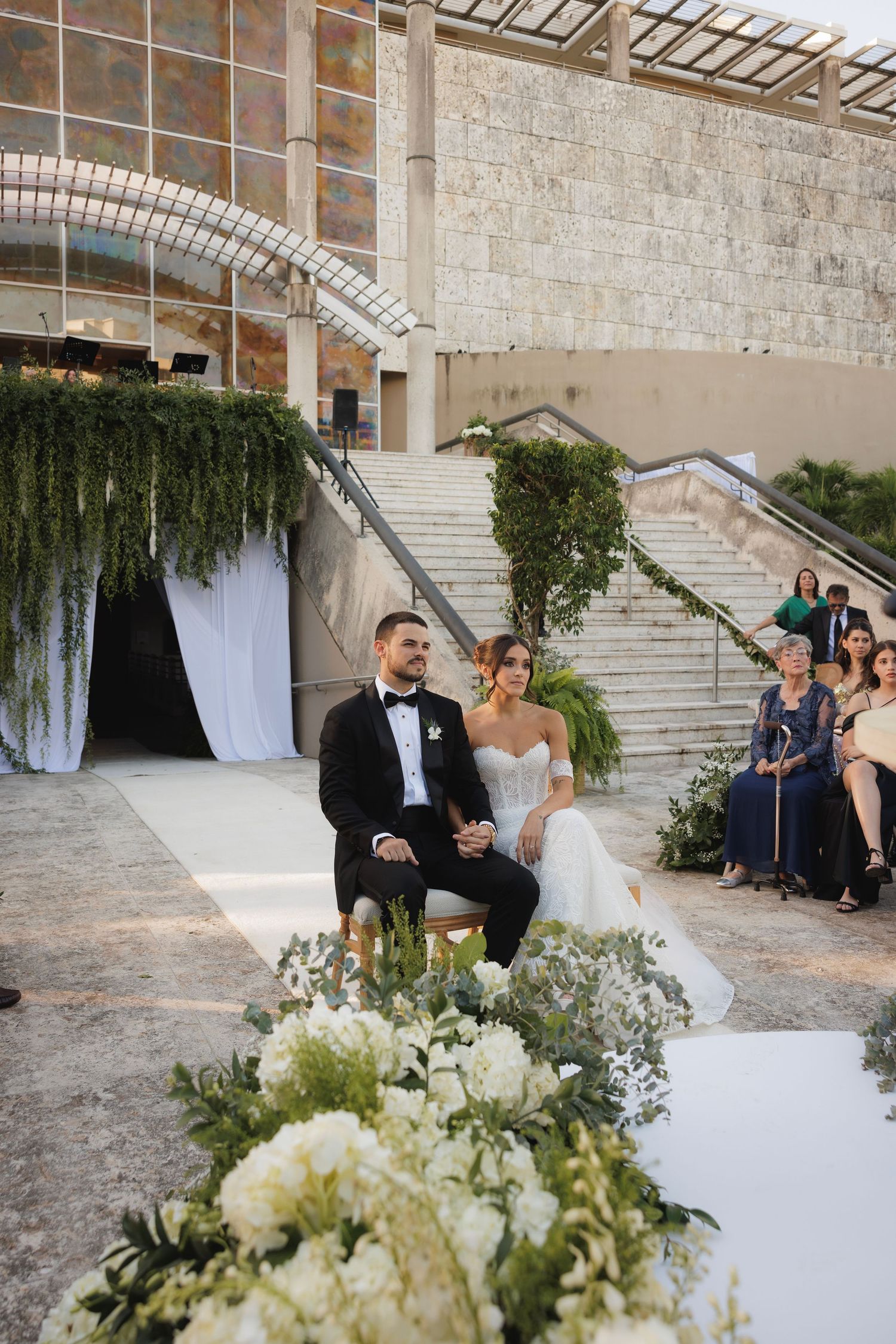 An intimate outdoor wedding ceremony setup with elegant white floral arrangements and draped fabric near grand stone steps.