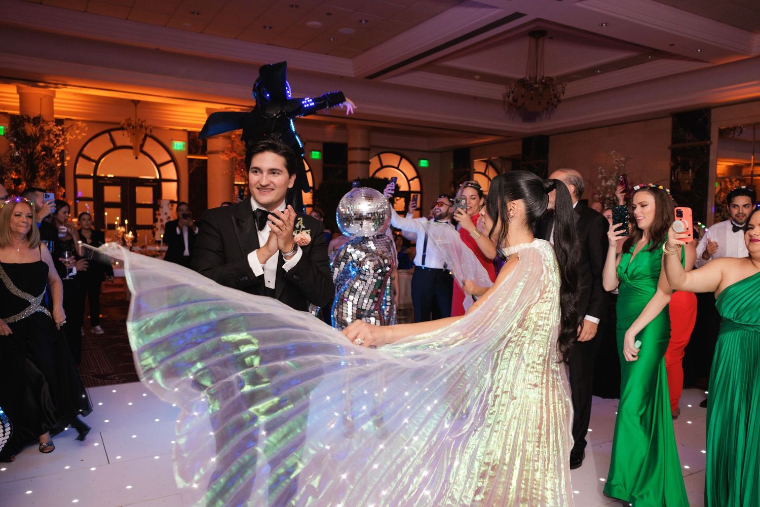 A wedding celebration features a dramatic flowing iridescent dress on the dance floor surrounded by guests in formal attire.