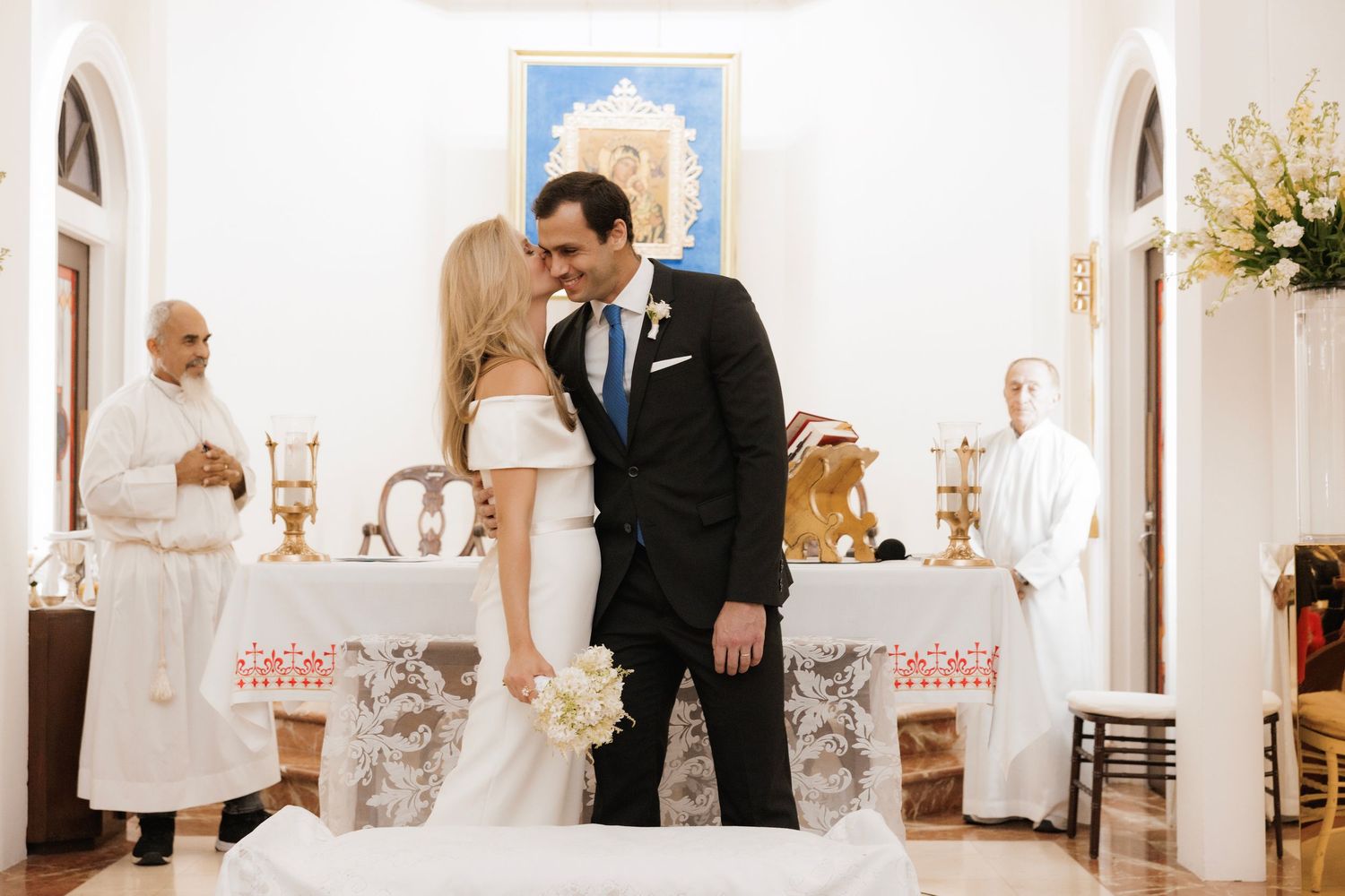 A couple shares a romantic moment at the altar with priests observing in an elegant white church setting.